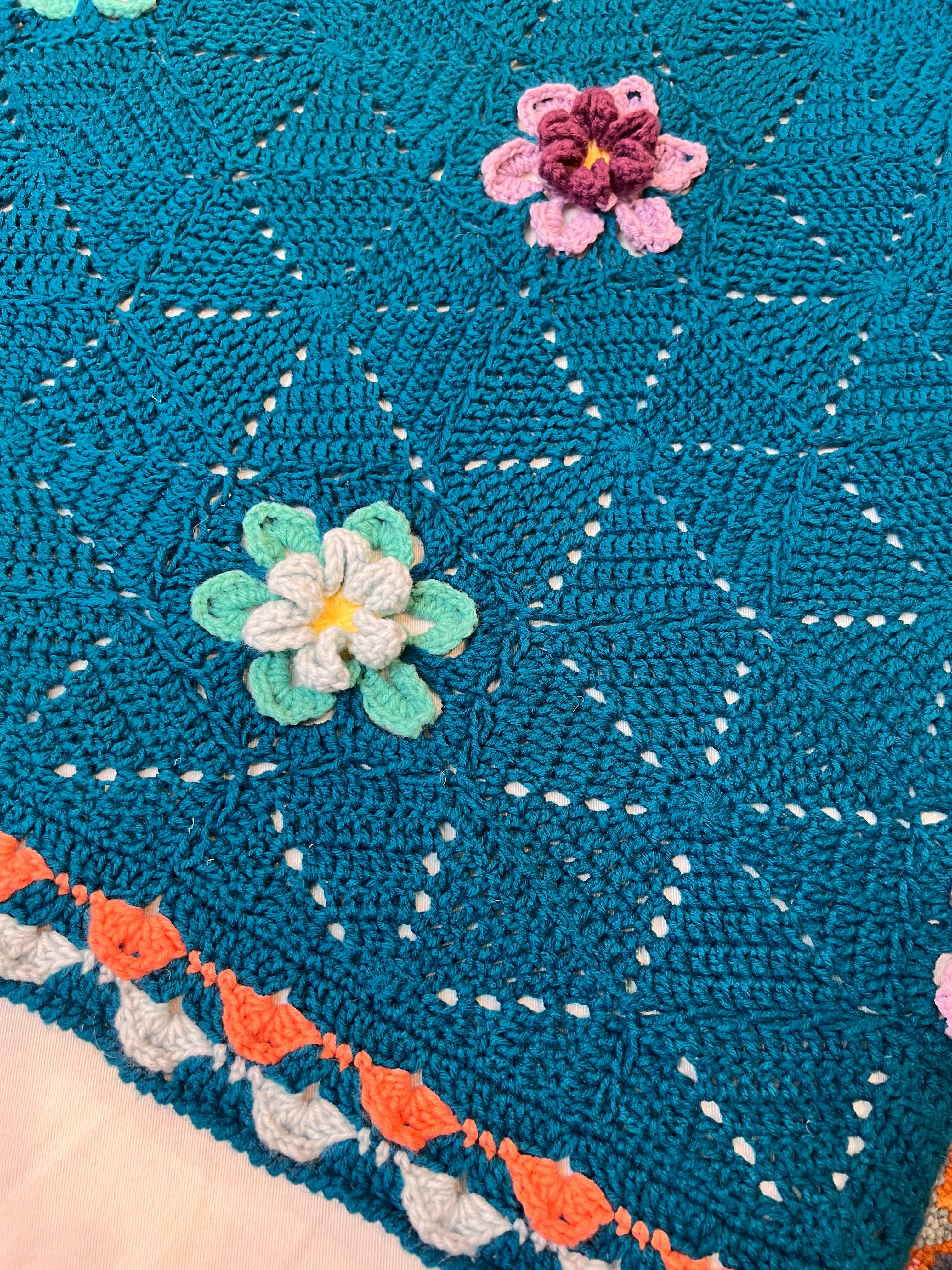 A close-up of a teal blanket crafted using the Wildflower Hexagon Motif Crochet Blanket Pattern, featuring colorful 3D flower motifs and a decorative scalloped border in orange, white, and teal, beautifully showcasing its intricate crochet motif design.