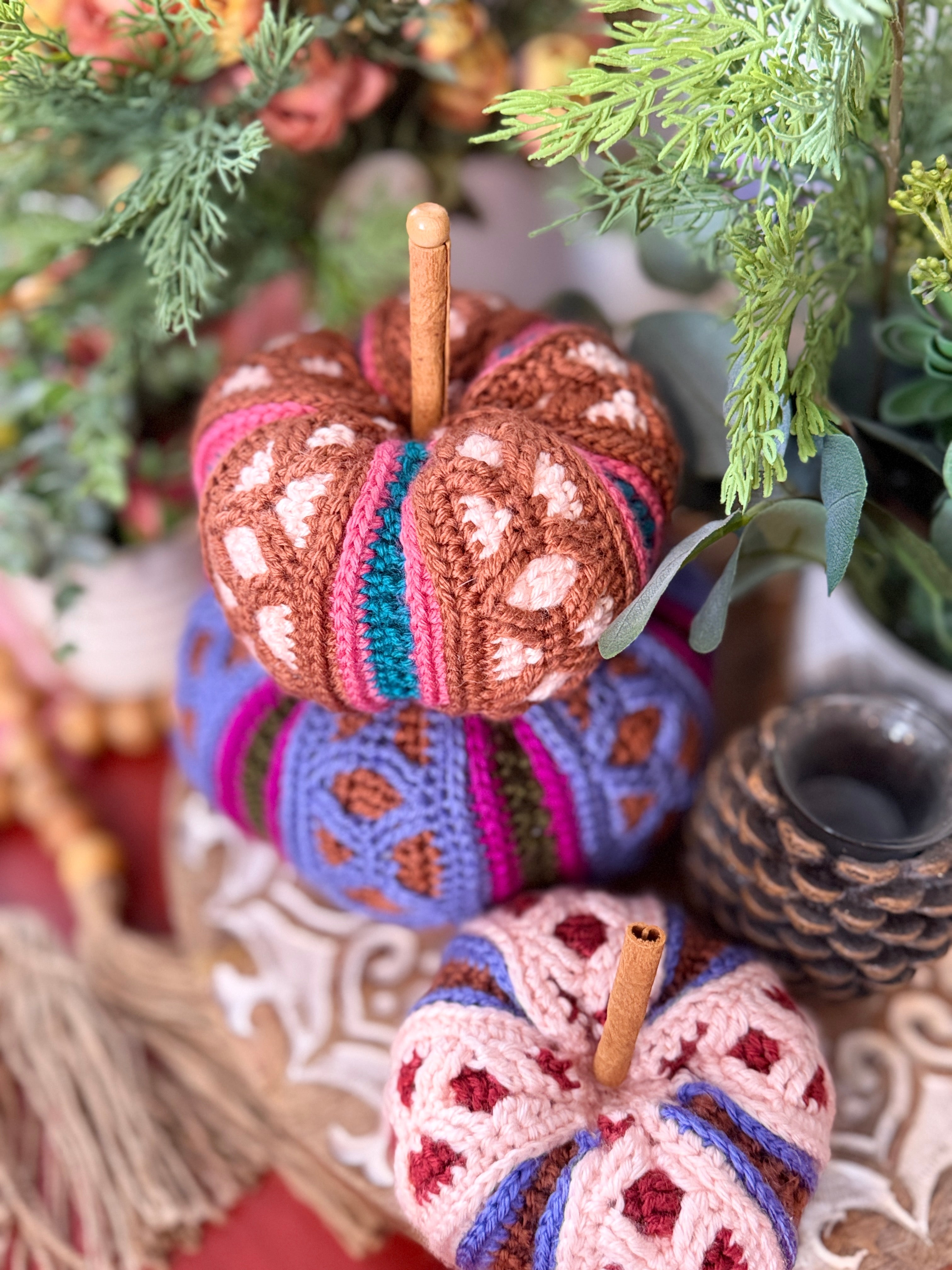 Three hand-crocheted decorative pumpkins stacked together in a fall-themed setting with green foliage, a candle, and a pinecone. The pumpkins feature intricate multicolor patterns of brown, pink, blue, purple, and red.