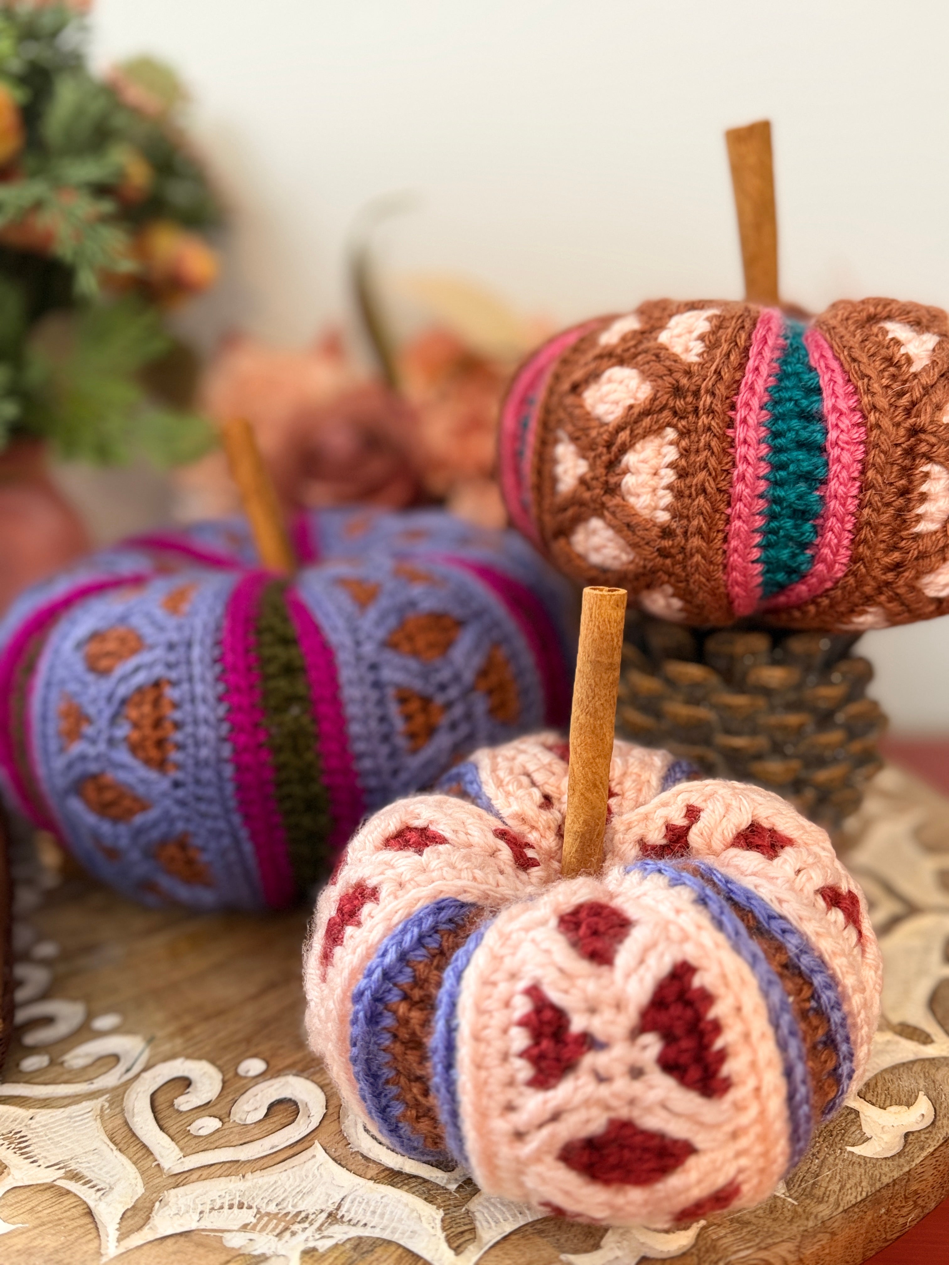 Three crocheted decorative pumpkins with wooden stems arranged on a wooden surface in a cozy indoor setting. The front pumpkin is cream with red and blue stripes and red floral motifs, the middle pumpkin is lavender with brown and magenta stripes, and the back pumpkin is brown with pink, teal, and cream stripes.