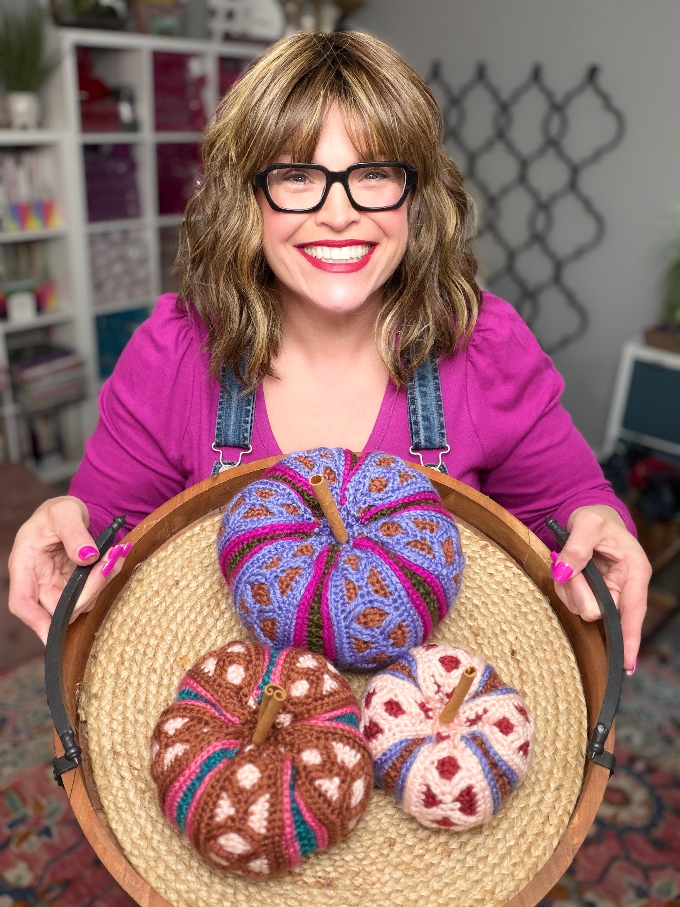 A smiling woman with glasses and a pink shirt holds a round wooden tray with three hand-knit crocheted pumpkins in purple, brown, and pink patterns. The background shows a cozy, styled room with shelves and home decor.