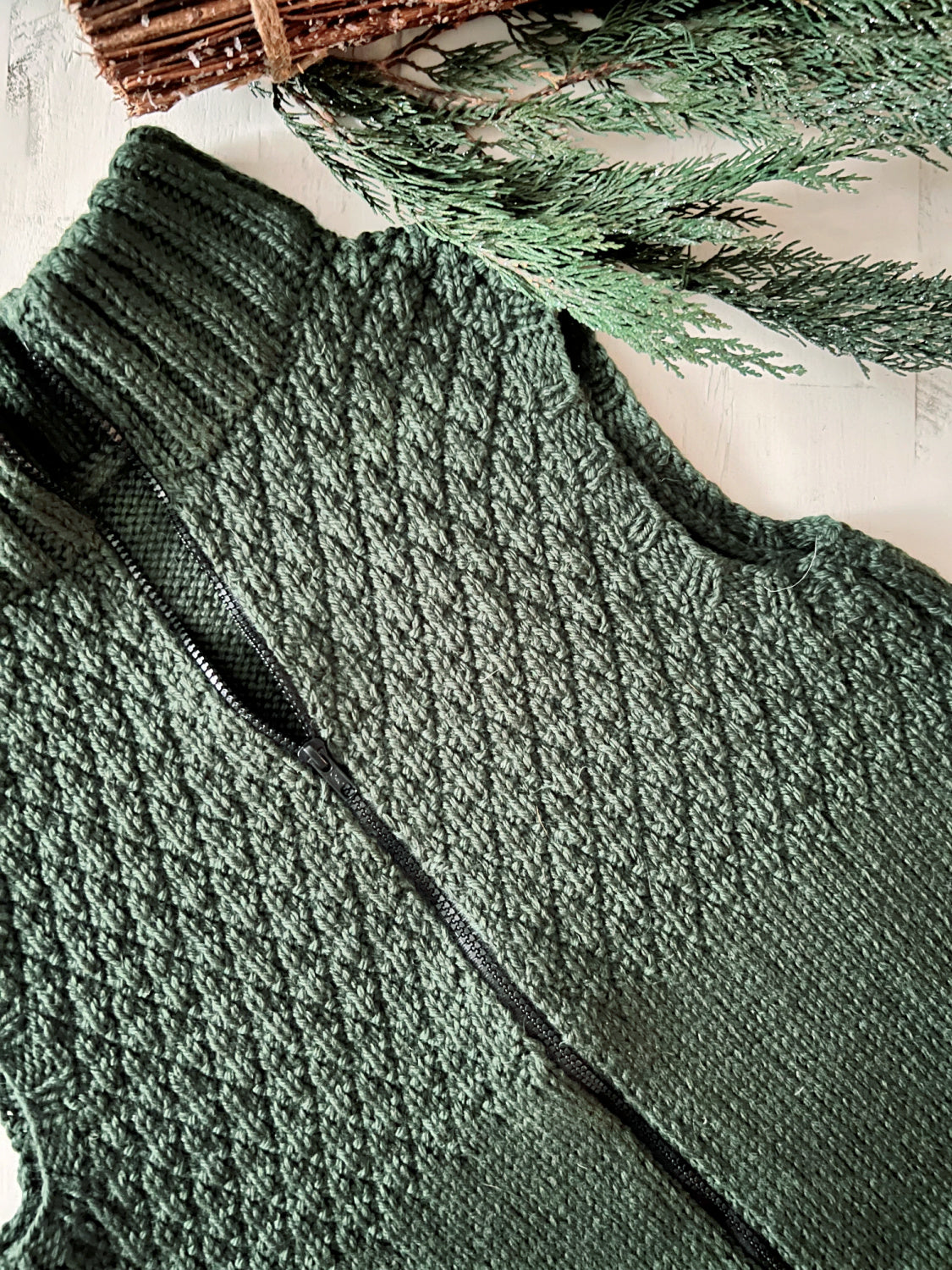 A close-up of the Harvey Knit Vest Pattern—a textured dark green zip-front knit vest with a black zipper, crafted in a seersucker stitch—lying on a light surface next to pine tree branches.