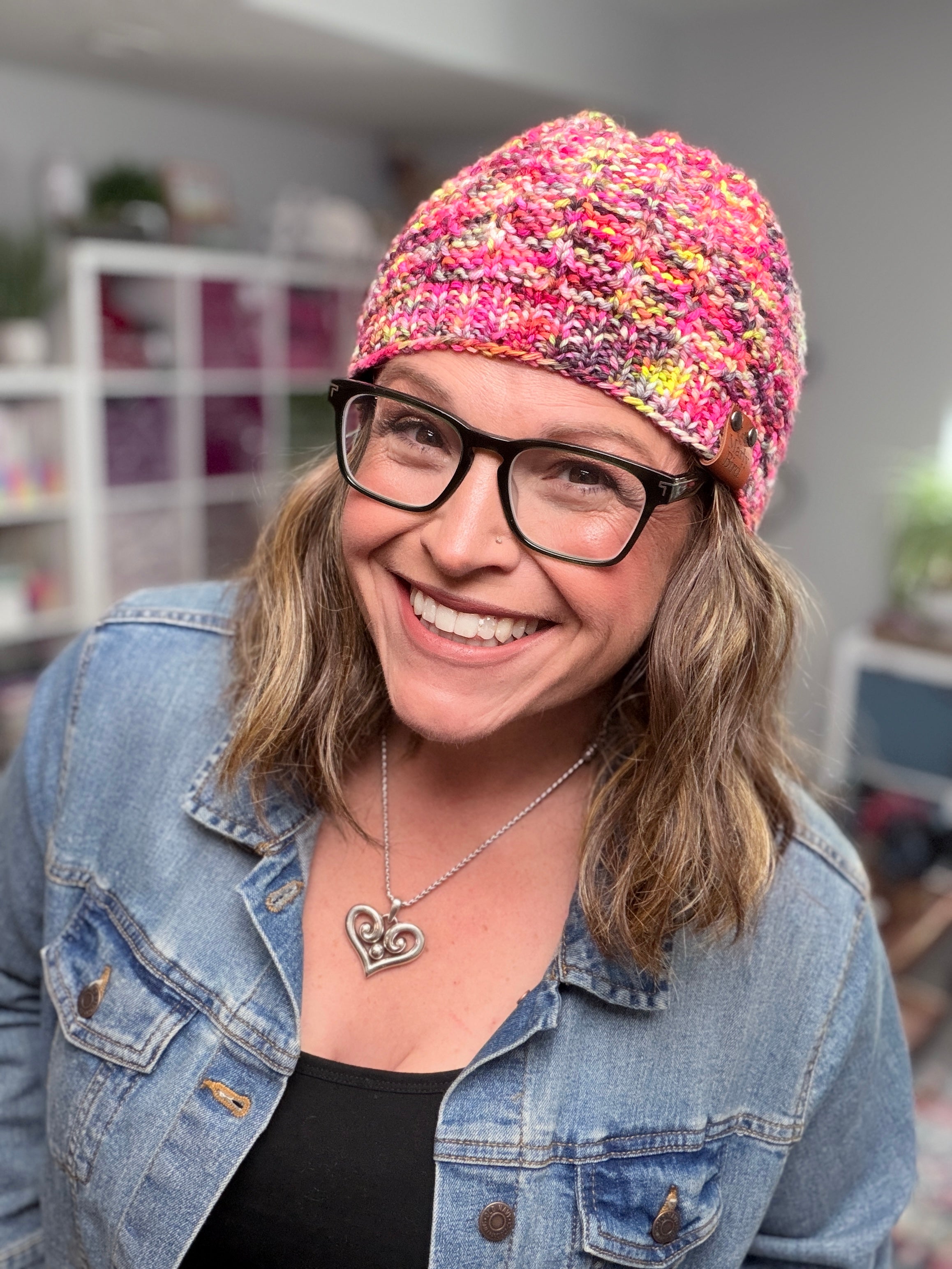 A smiling woman in glasses models the Jubilee Knit Hat, featuring lifted knit ribbing, as she stands indoors wearing a denim jacket and heart-shaped necklace, with blurred shelves and plants in the background.
