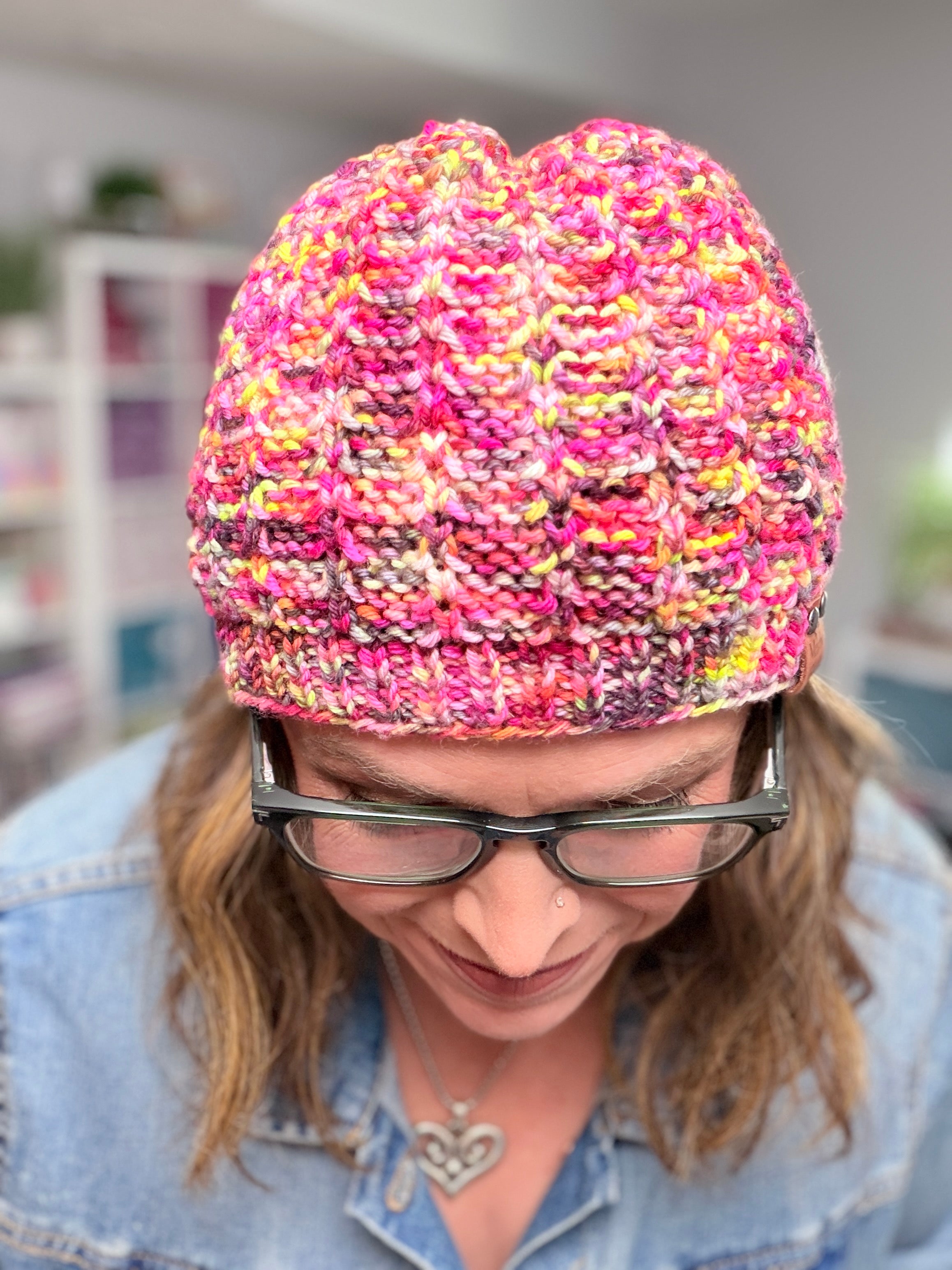 A person wearing glasses and the Jubilee Knit Hat—a colorful pink design crafted with multicolor yarn—looks down, smiling. They have long hair and are dressed in a blue denim shirt. The softly blurred background features shelves and plants.