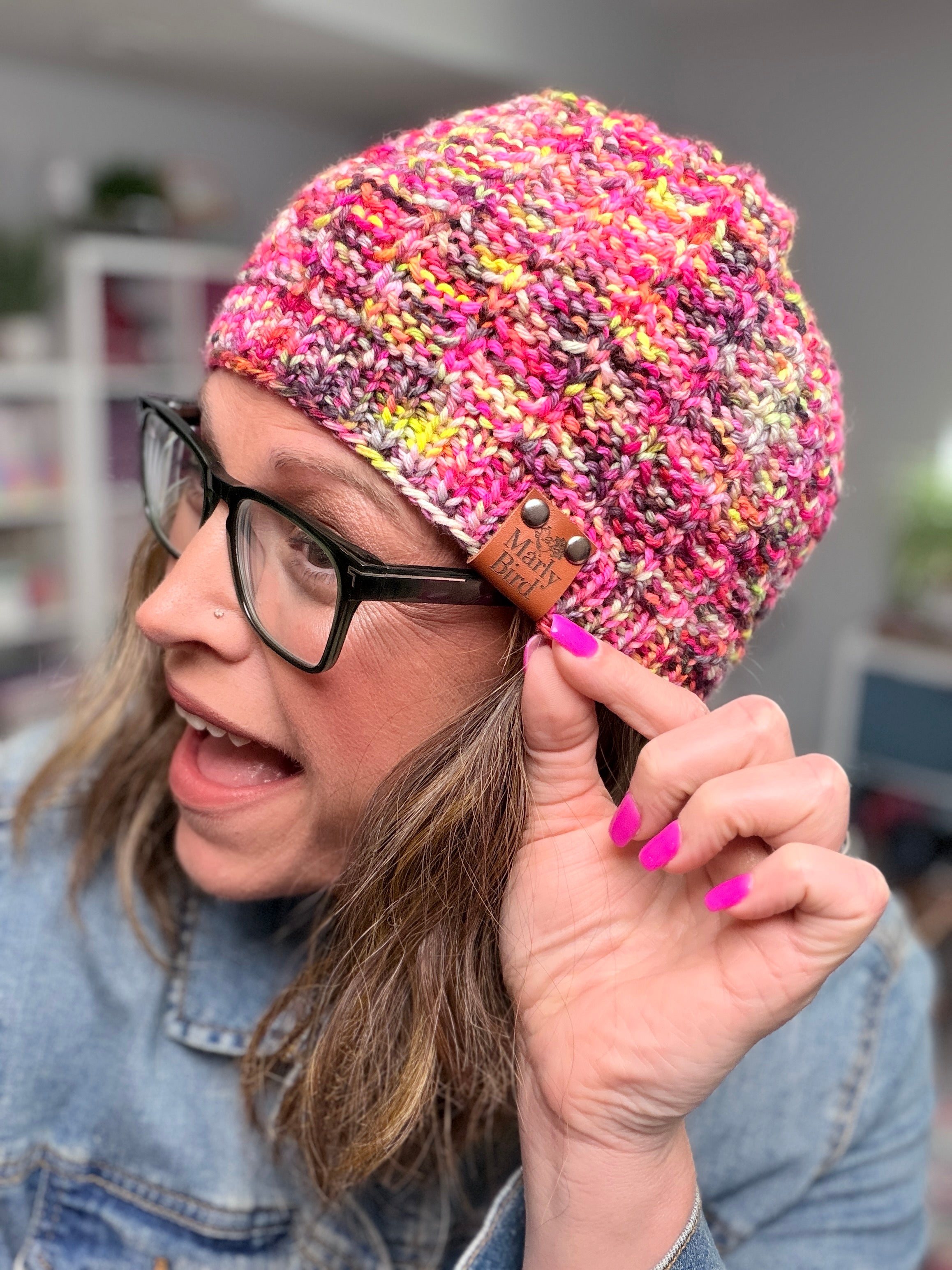 A person with glasses and pink nails smiles while showing off a vibrant beanie, handmade from the Jubilee Knit Hat Pattern with multicolor yarn and adorned with a “Makers Gonna Make” tag. The background is softly blurred.