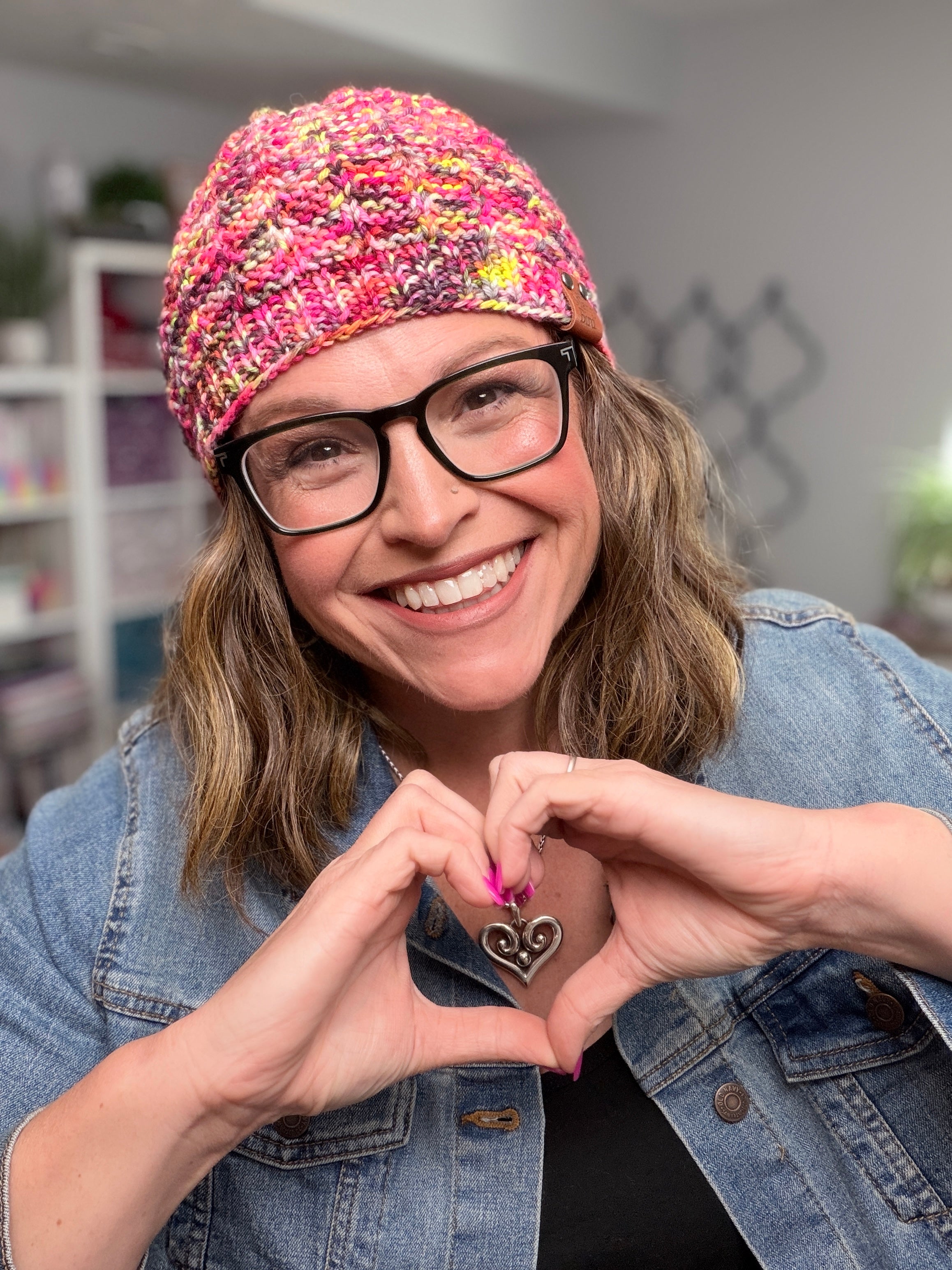 A smiling person wearing glasses and the Jubilee Knit Hat, featuring lifted knit ribbing in a pink shade, makes a heart shape with their hands to showcase a heart-shaped pendant. They are dressed in a denim jacket, with a bookshelf and decor visible in the background.