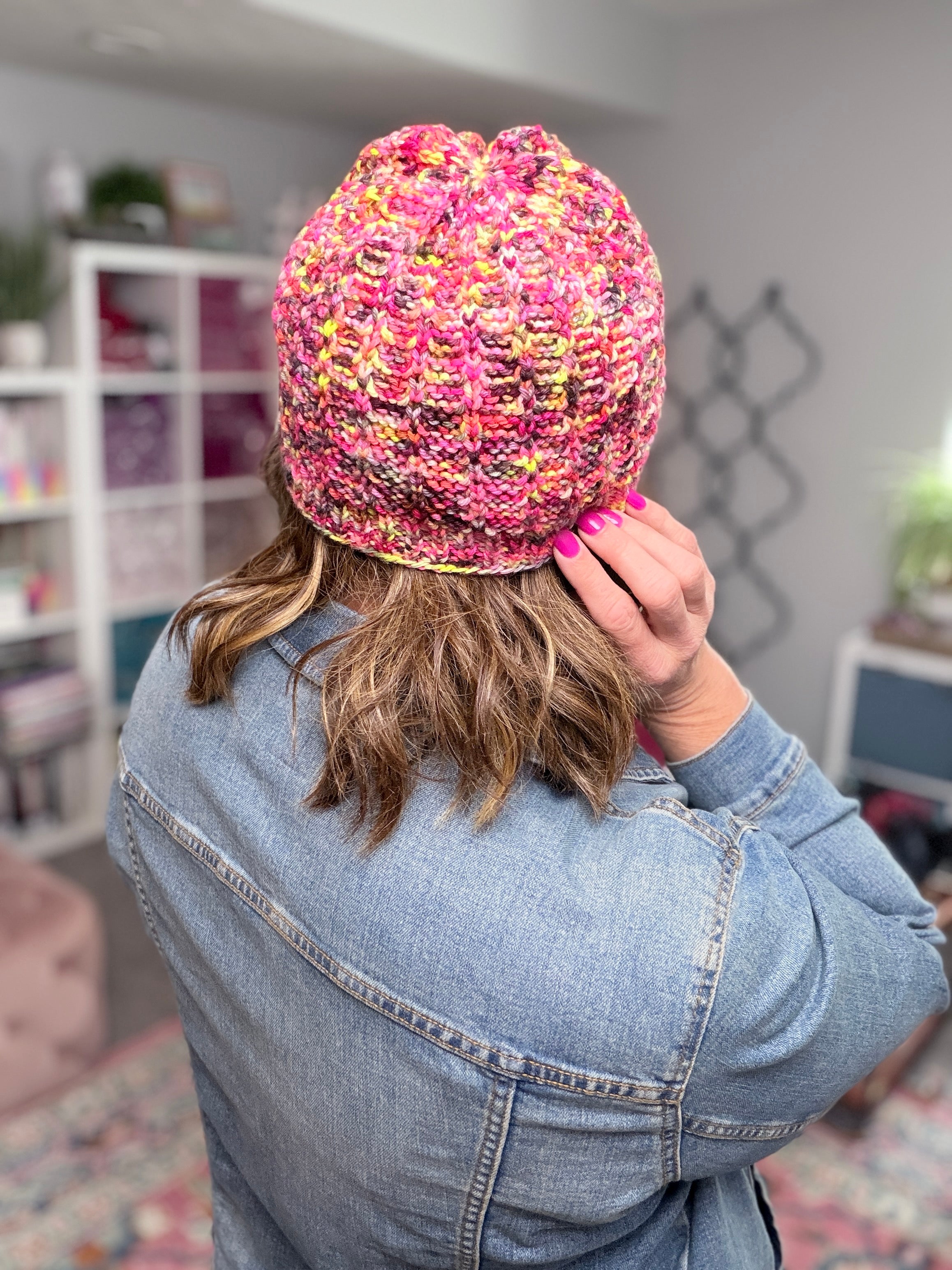 A person wearing the Jubilee Knit Hat, in pink, yellow, and purple, stands with their back to the camera. Their shoulder-length hair and denim jacket are visible as they hold the hat—showing off pink nail polish—with a hand raised to their head.