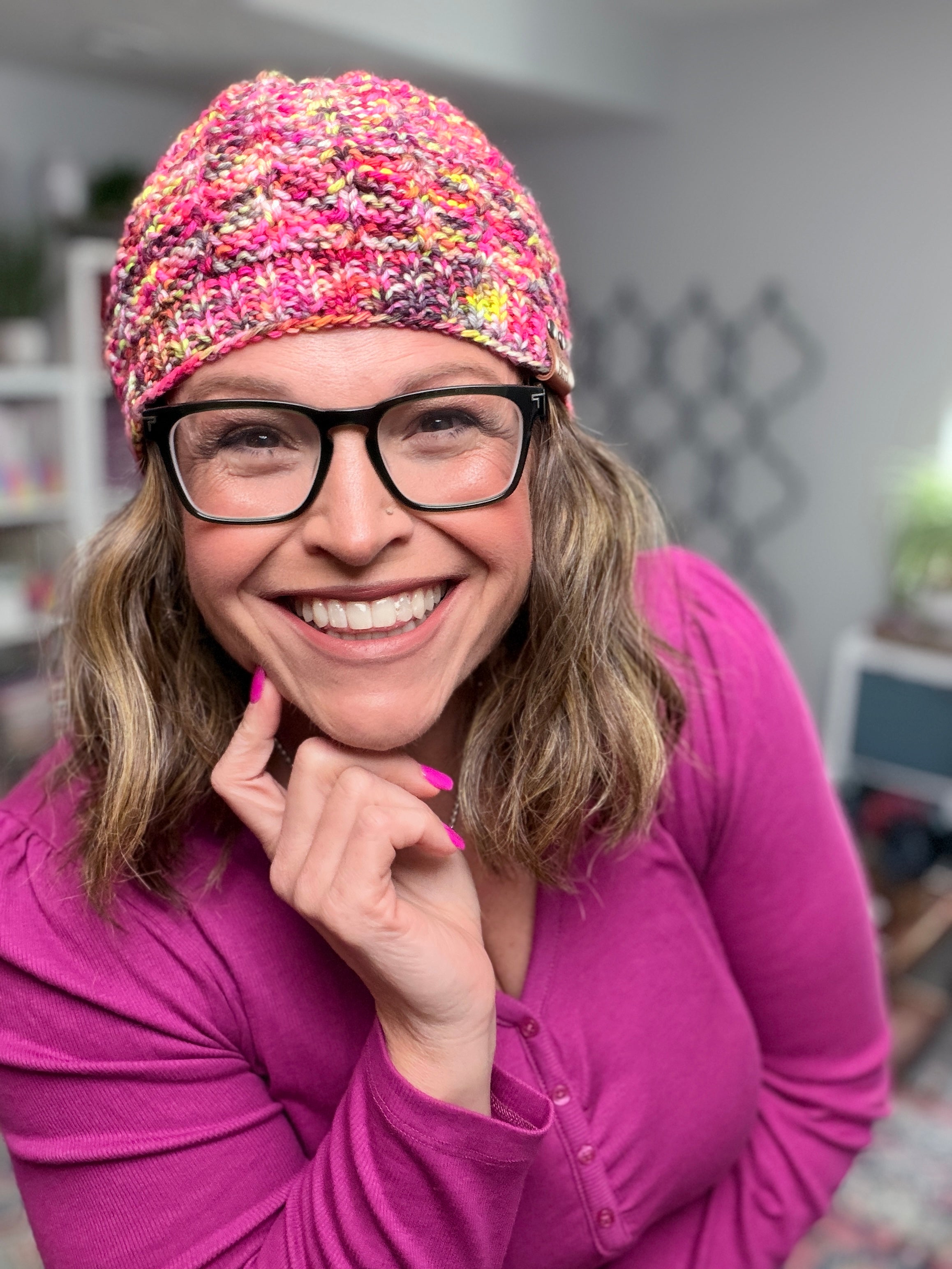 A smiling woman wearing glasses and a pink shirt poses indoors, touching her chin with one hand, while modeling the Jubilee Knit Hat—a colorful hat made from multicolor yarn. Blurred shelves and home decor appear in the background.