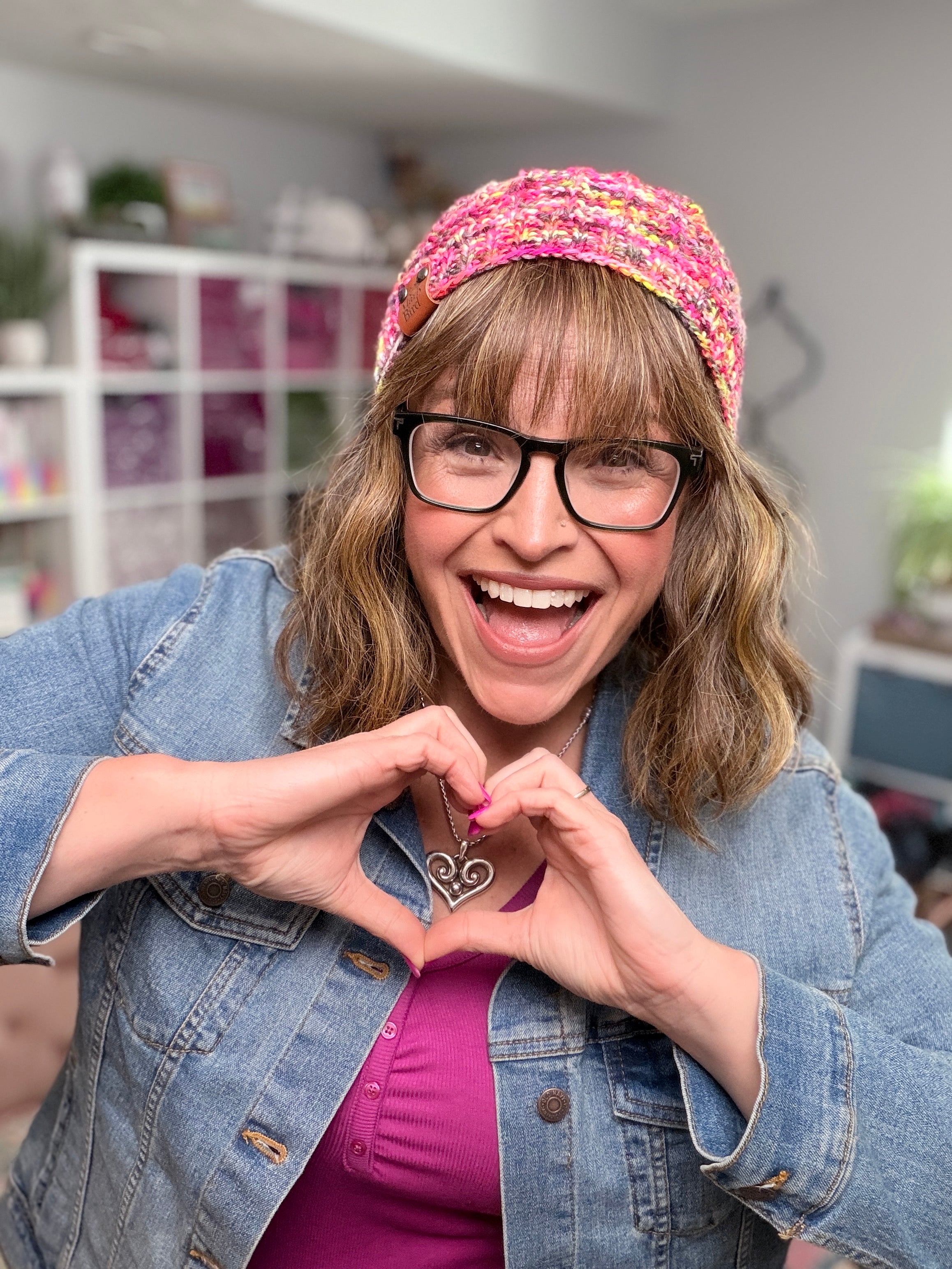 A smiling woman wearing glasses and a Jubilee Knit Hat—featuring colorful yarn and lifted knit ribbing—pairs it with a denim jacket as she makes a heart shape with her hands. She stands indoors, with shelves and plants softly blurred in the background.