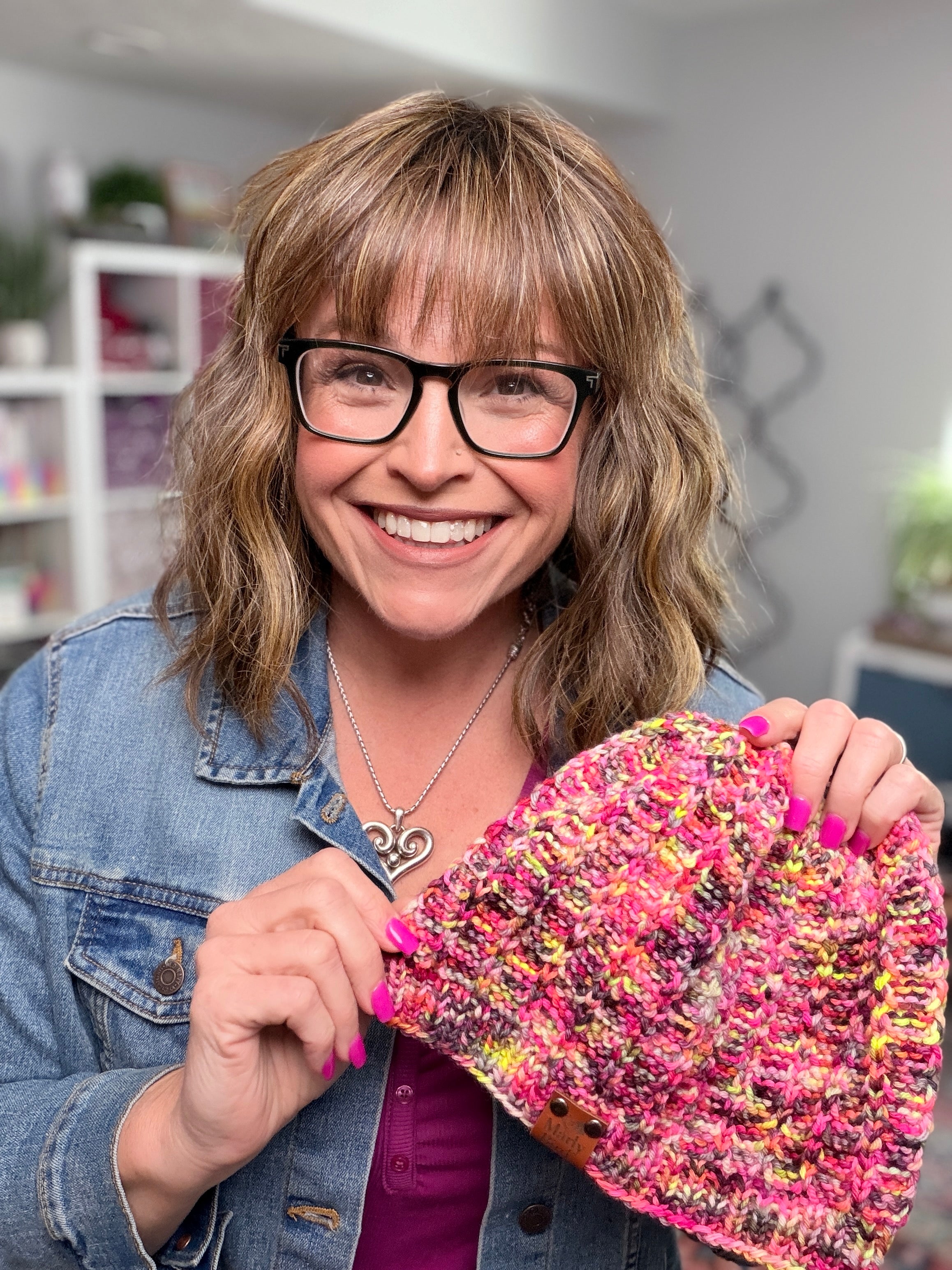 A smiling person with glasses and wavy hair, dressed in a denim jacket, holds up a Jubilee Knit Hat made from vibrant multicolor yarn. The softly blurred background features shelves and plants.