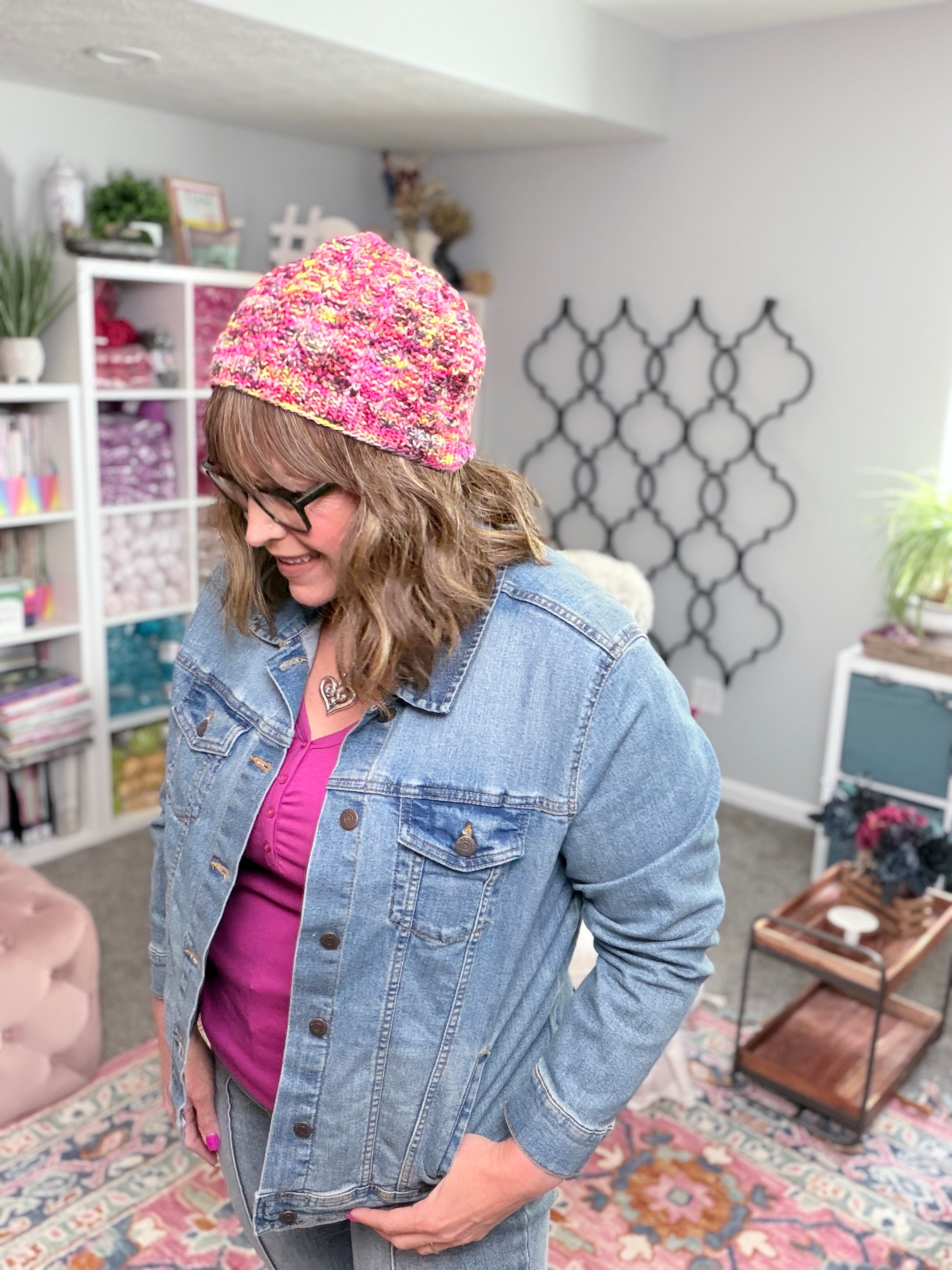 A woman wearing glasses and a handmade Jubilee Knit Hat, featuring lifted knit ribbing in pink yarn, pairs it with a denim jacket as she stands in a cozy, bright room adorned with bookshelves, plants, and colorful decor.
