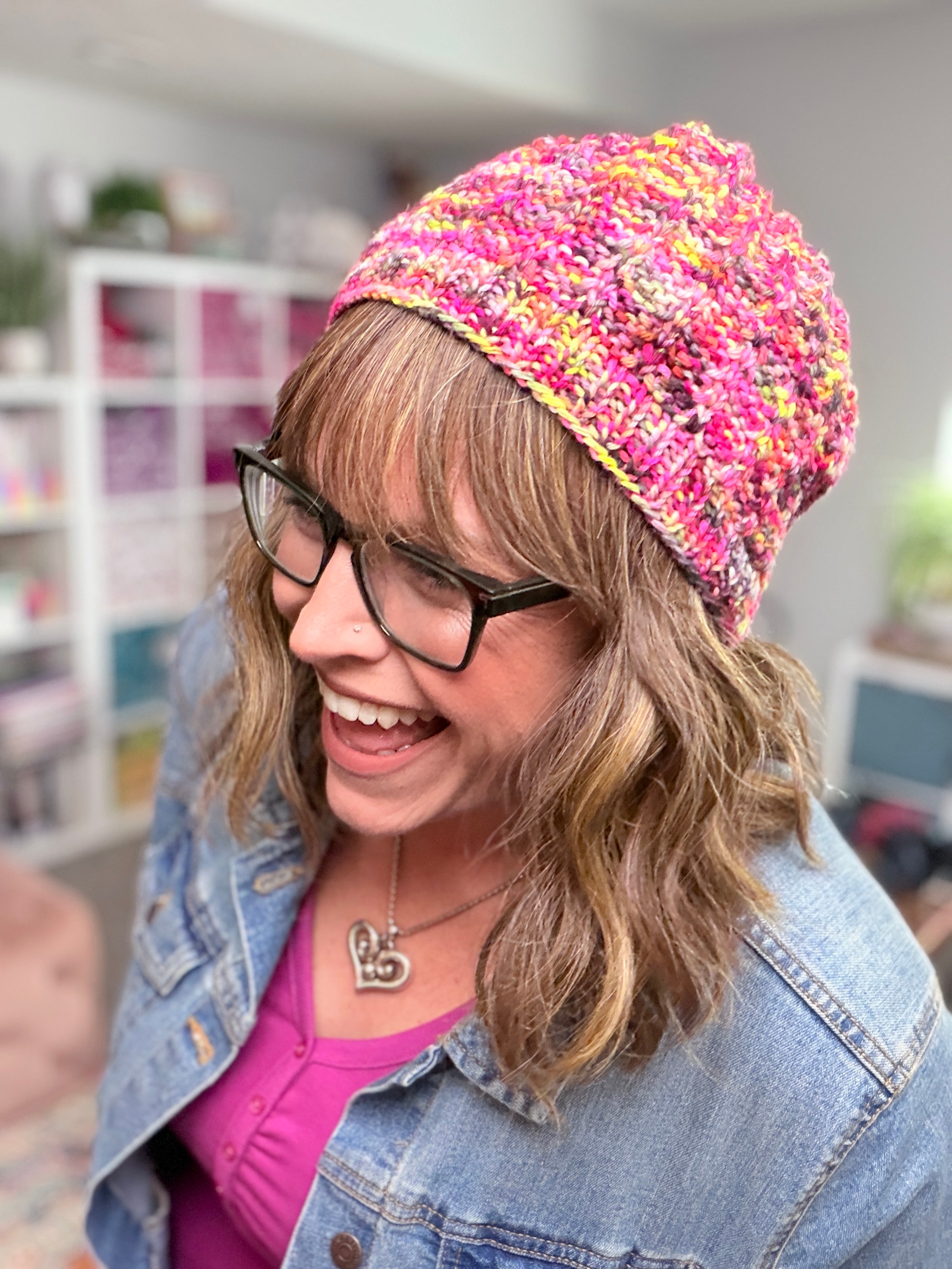 A smiling woman with wavy hair and glasses showcases the Jubilee Knit Hat Pattern—a colorful crocheted hat made from multicolor yarn—paired with a heart necklace, pink top, and denim jacket, standing indoors with shelves and plants in the background.