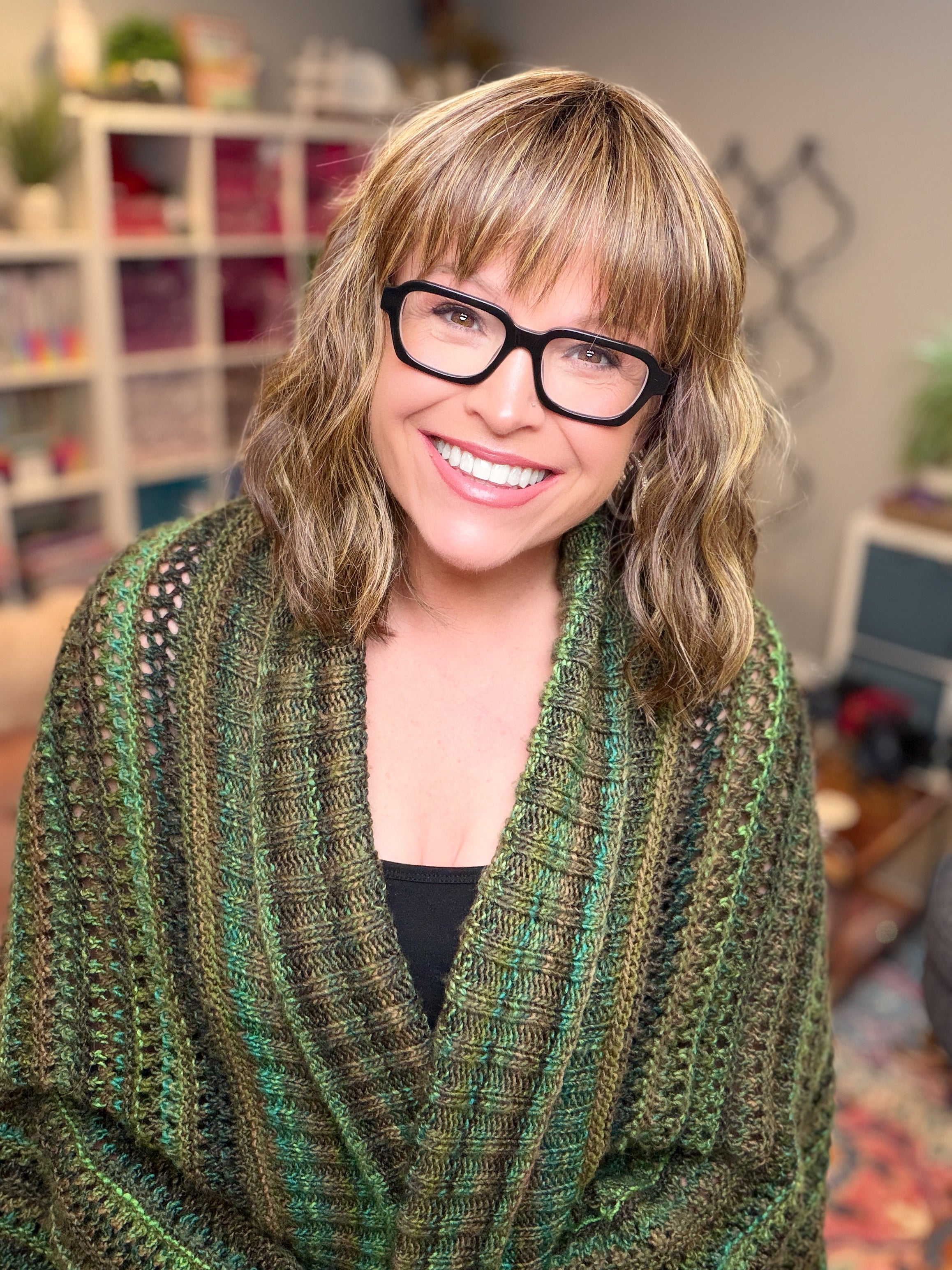A smiling person with wavy hair and glasses is wearing an oversized cocoon cardigan made with the Know Your Worth Knit Cocoon Cardigan Pattern, featuring a textured knit design. They are indoors, surrounded by shelves of colorful yarn and craft supplies in the background.