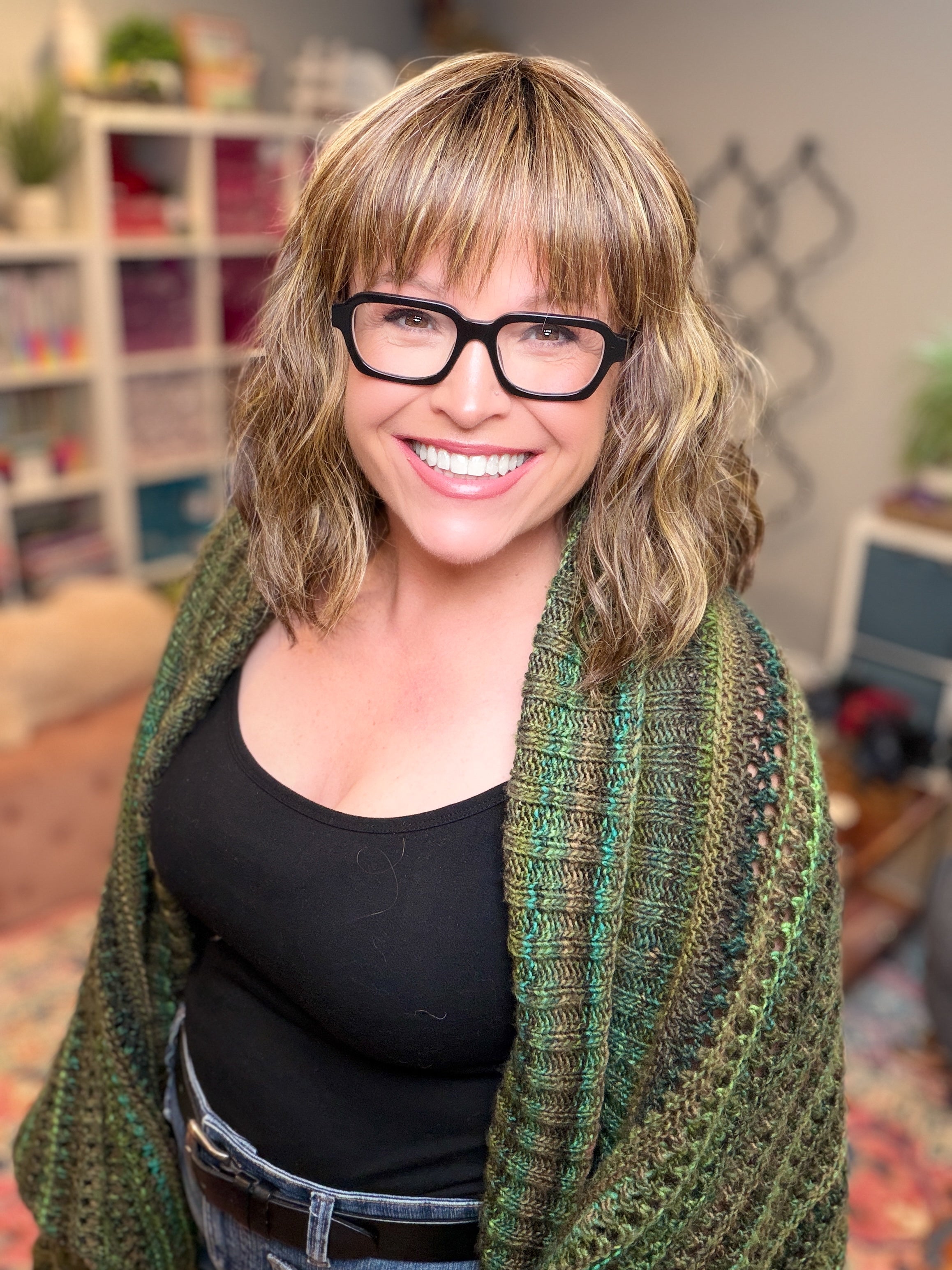 A smiling person with wavy, blond hair and glasses stands in a cozy, colorful room filled with shelves and plants, wearing a black top layered with the Know Your Worth Knit Cocoon Cardigan.