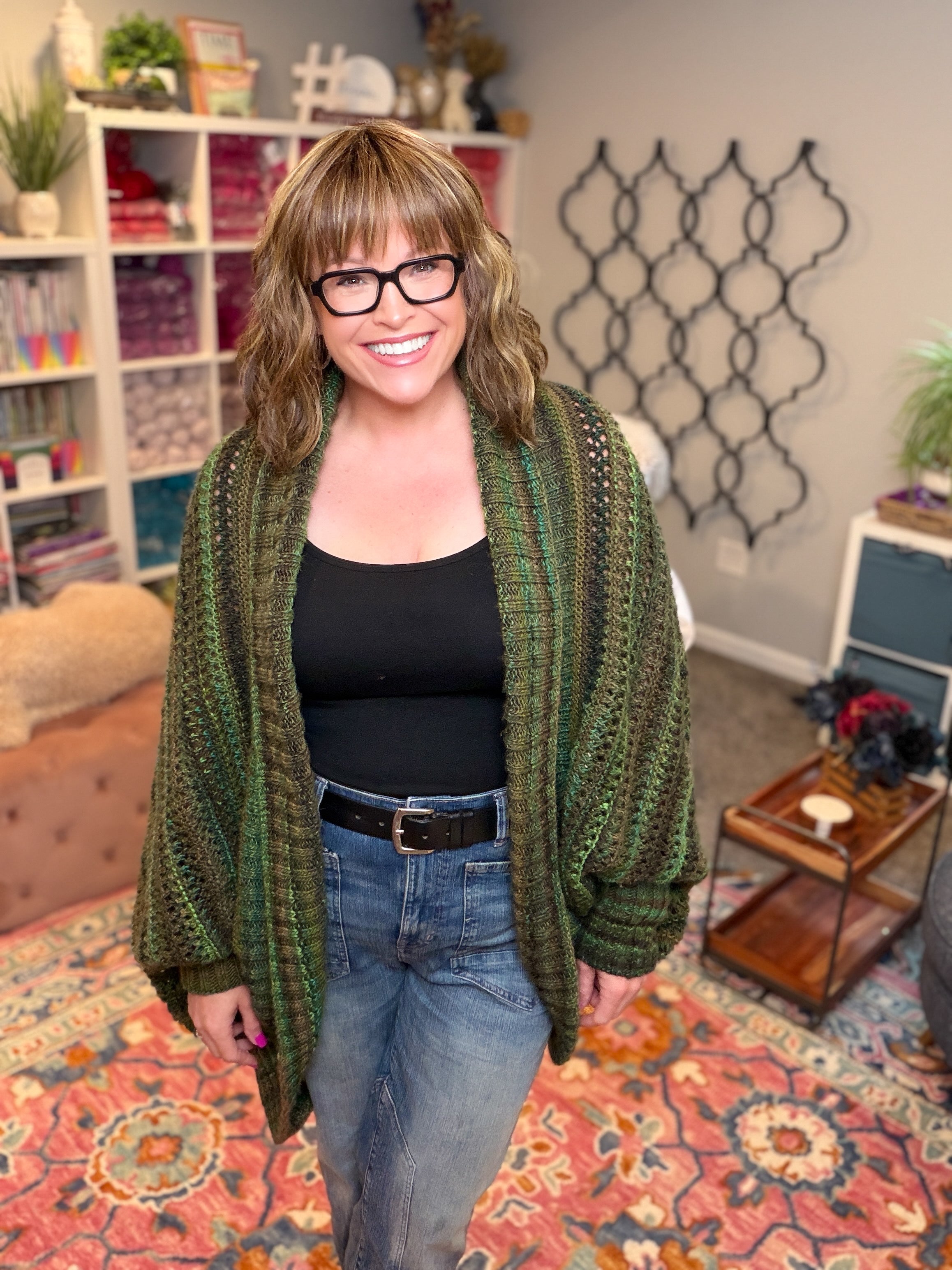 A smiling woman with glasses wears the Know Your Worth Knit Cocoon Cardigan, which features a textured knit pattern in green. She pairs it with a black top and jeans while standing in a cozy room adorned with colorful yarn shelves, a patterned rug, and decorative wall art in the background.