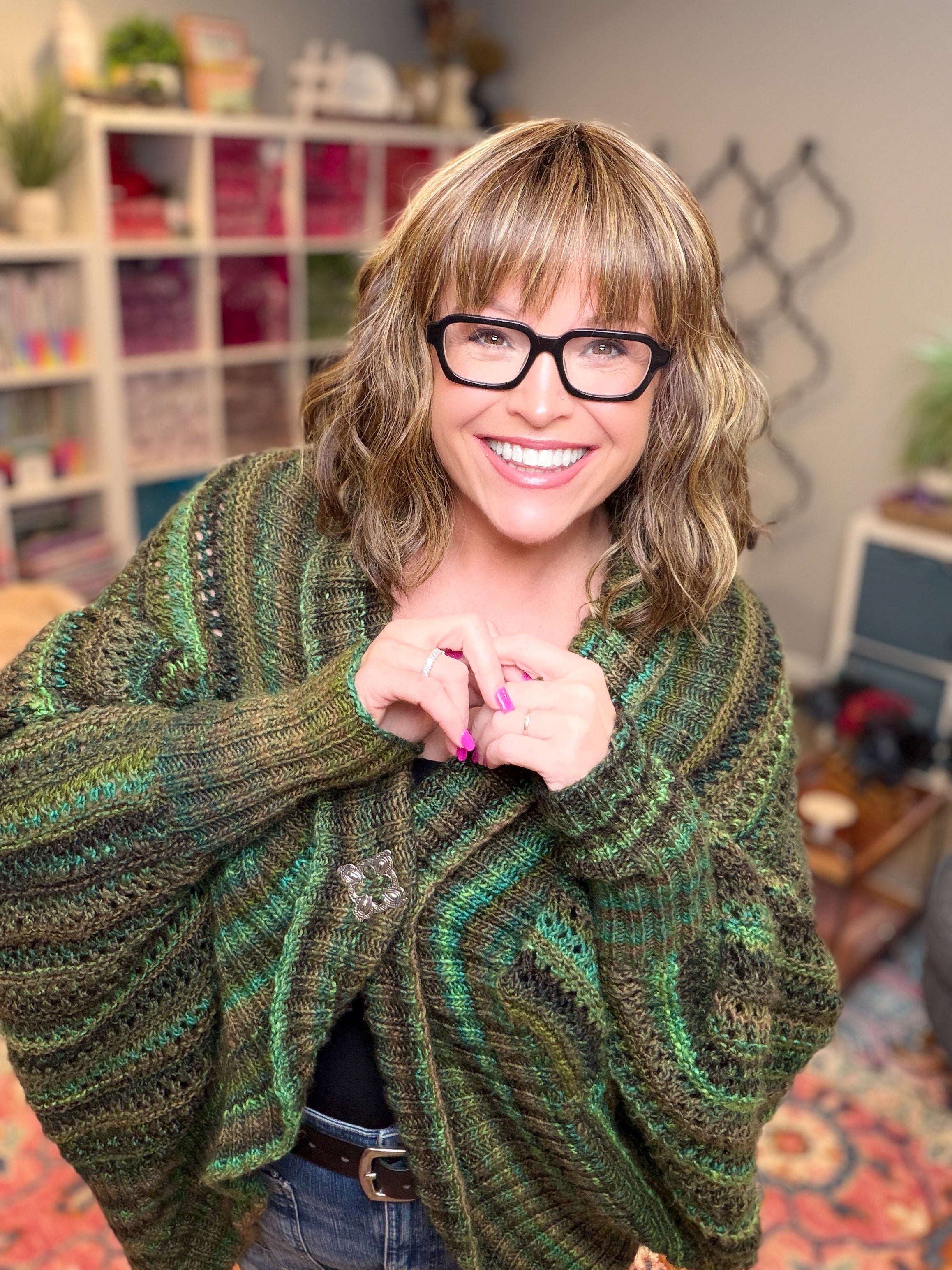 A smiling woman with glasses and shoulder-length hair models the Know Your Worth Knit Cocoon Cardigan Pattern in a vibrant green, featuring an oversized fit and textured knit design. She stands indoors with colorful shelves and decor in the background, hands together to show off her pink nail polish.