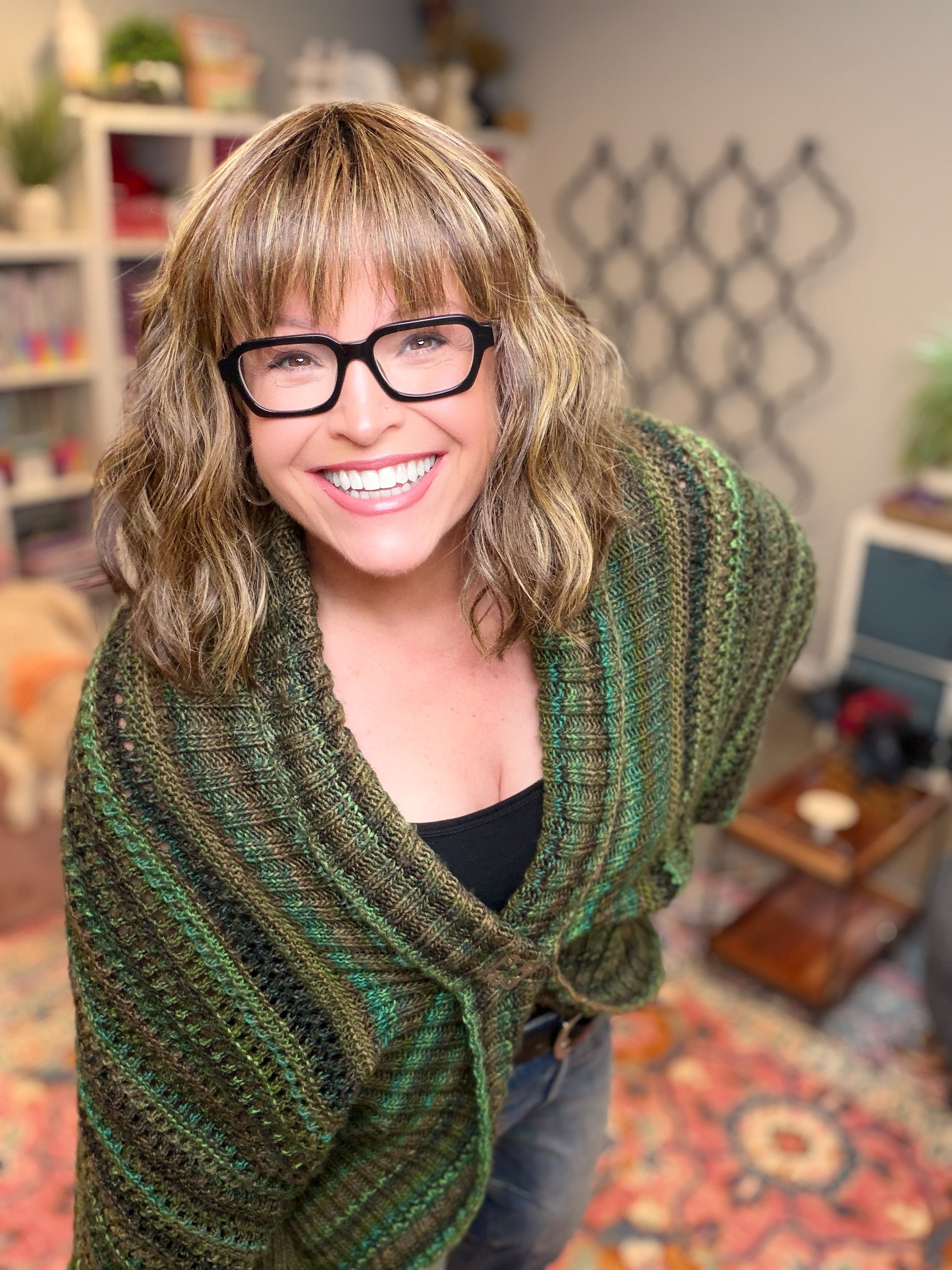 A smiling person with glasses and wavy hair wears the Know Your Worth Knit Cocoon Cardigan in a soft green shade, standing in a cozy, colorful room filled with shelves, plants, and patterned carpet in the background.