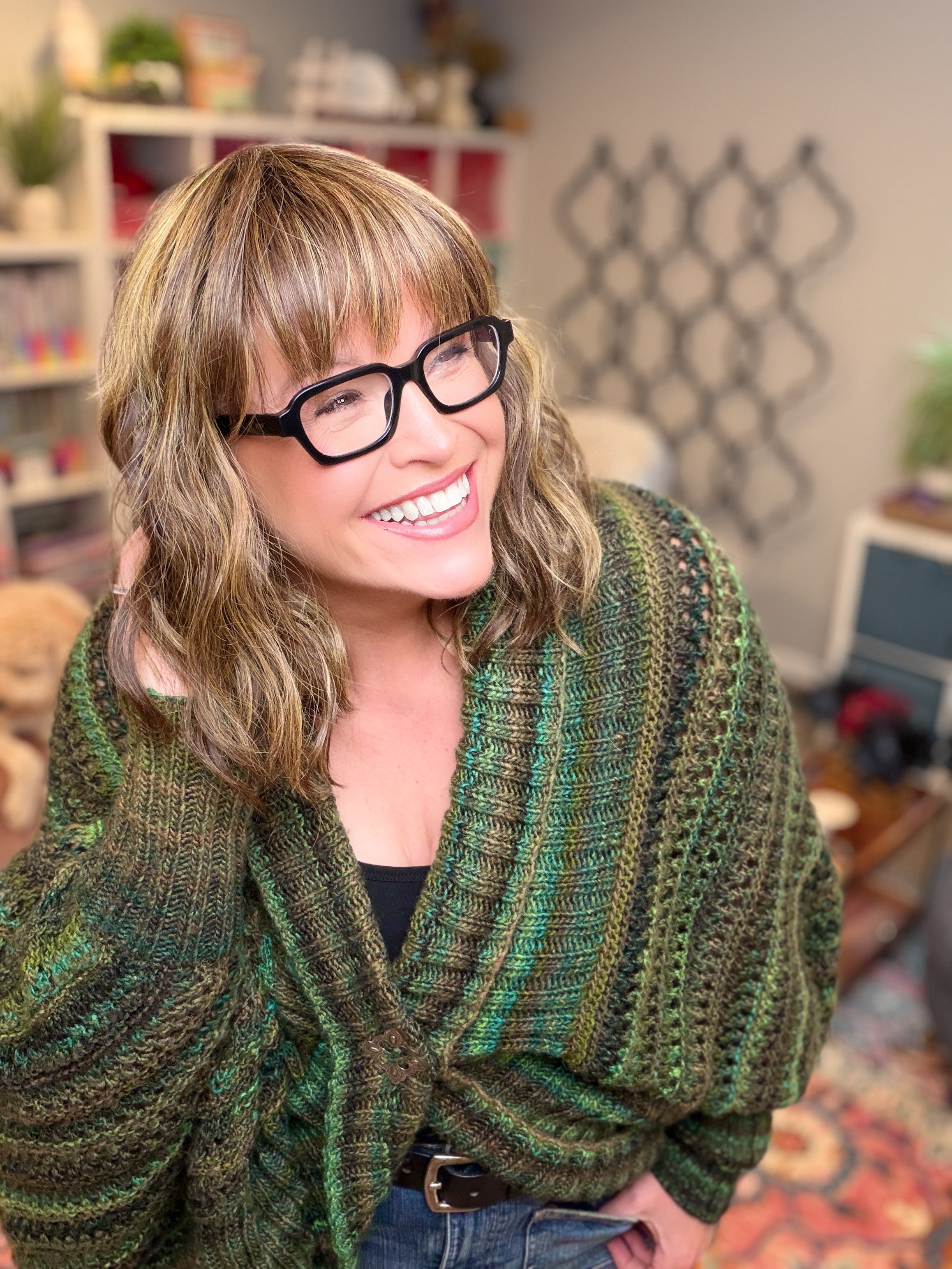 A woman with blond hair and glasses smiles while wearing the Know Your Worth Knit Cocoon Cardigan. She stands in a cozy room filled with shelves, plants, and books, the textured knit pattern of her cardigan enhancing the inviting atmosphere.