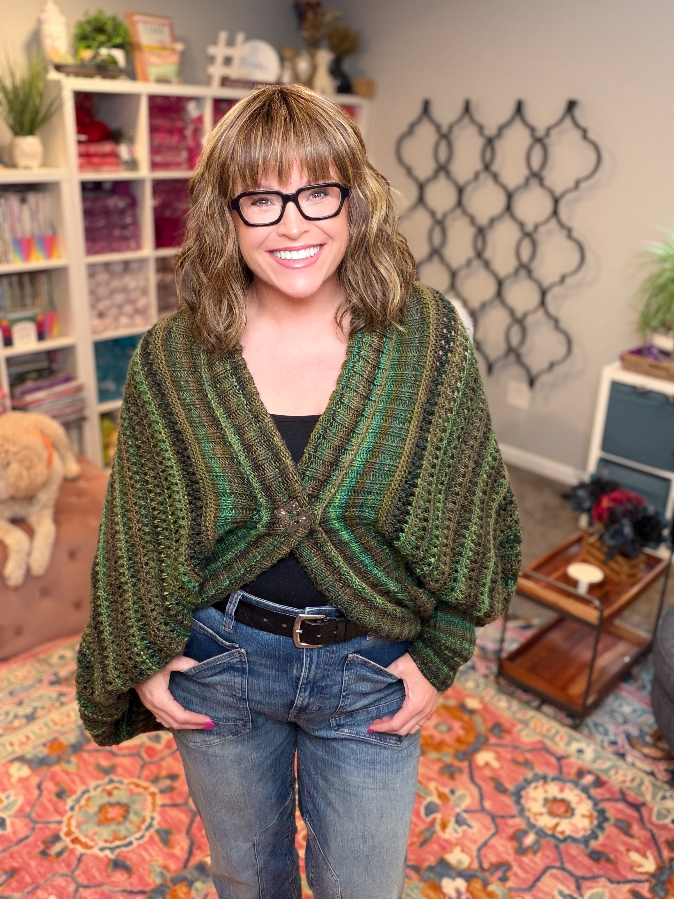 A smiling woman with shoulder-length hair and glasses models the Know Your Worth Knit Cocoon Cardigan Pattern in green stripes, paired with jeans, as she stands in a colorful, cozy room filled with yarn shelves, a patterned rug, and decorative wall accents.