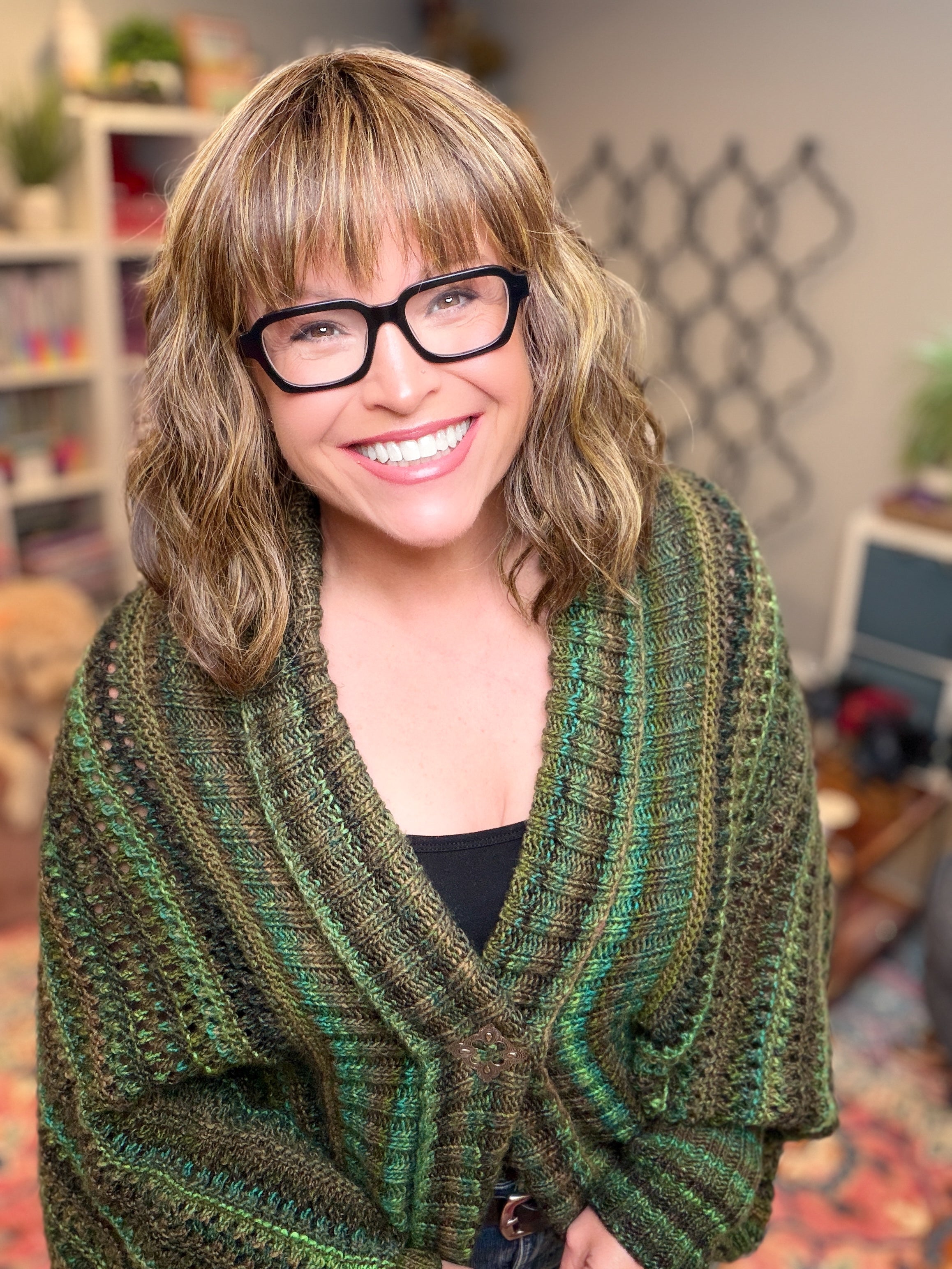 A smiling woman with wavy, shoulder-length hair and glasses wears the Know Your Worth Knit Cocoon Cardigan, featuring a textured knit pattern in green. She stands indoors with bookshelves and plants in the background.