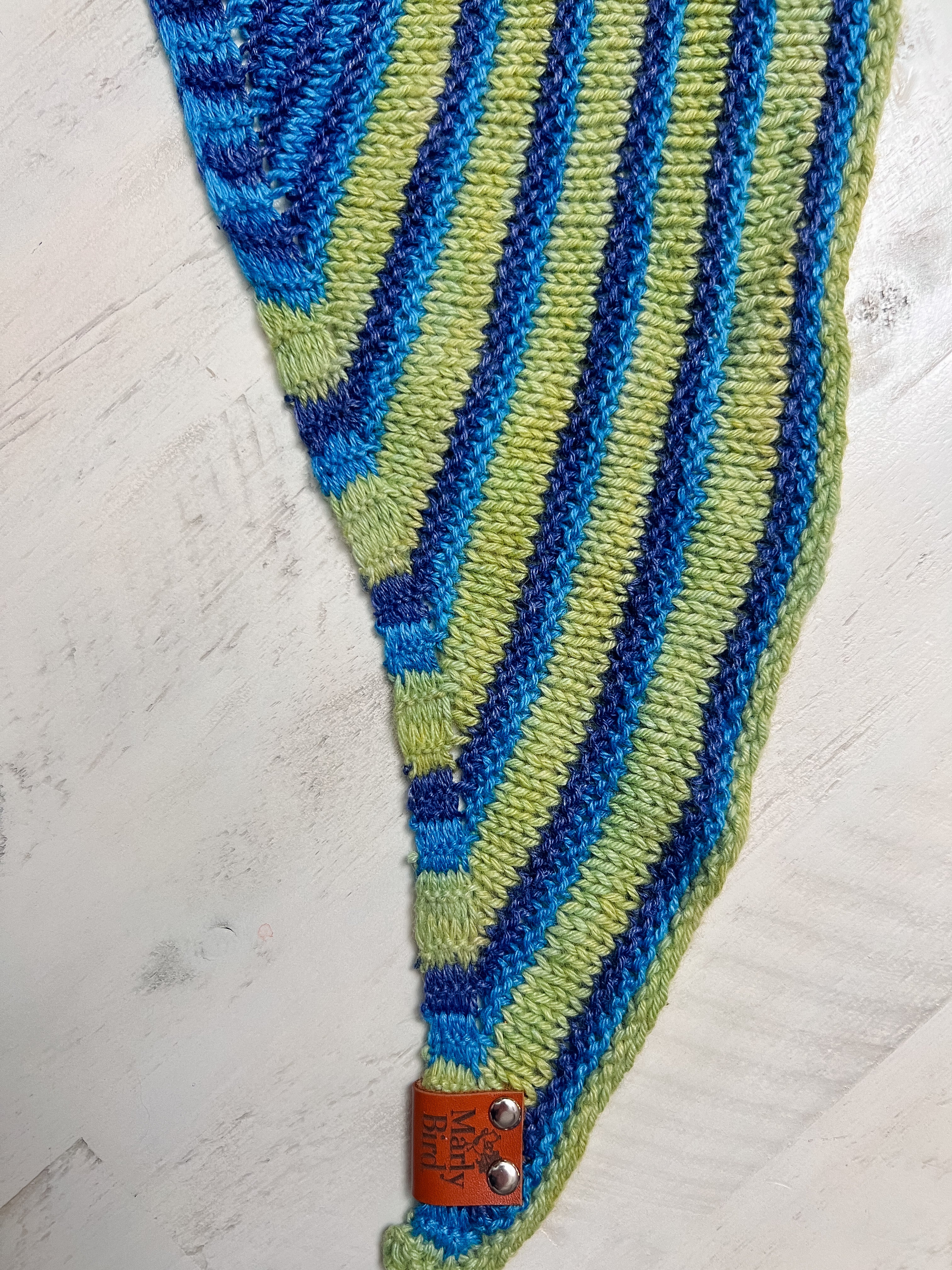 Green and blue striped fabric with a leather tag on a light gray background. Make It Mine Knit Shawl triangle shawl pattern garter stitch striped knit wrap