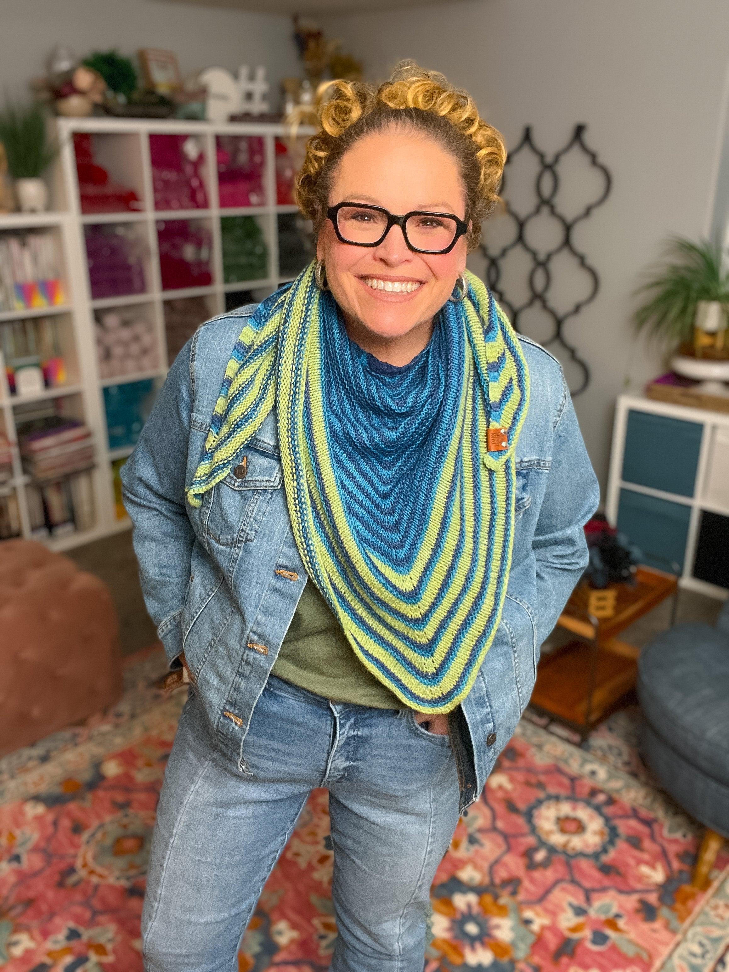 Person wearing a blue and green striped scarf in a room with a bookshelf and decorative items. Make It Mine Knit Shawl triangle shawl pattern garter stitch striped knit wrap