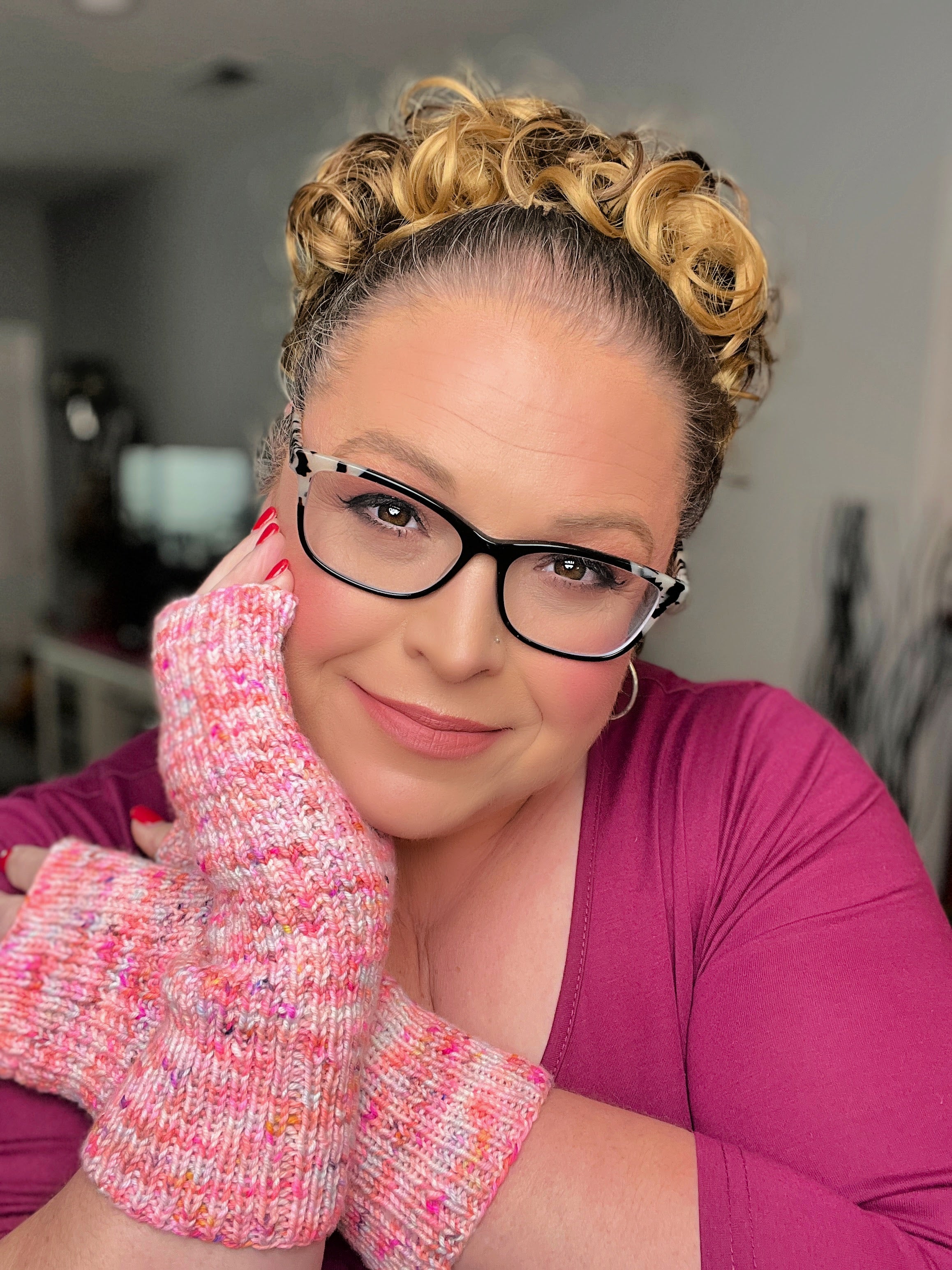 Pink textured fingerless mittens worn with wrists crossed, showing the ribbed cuffs and thumb shaping against a magenta top.