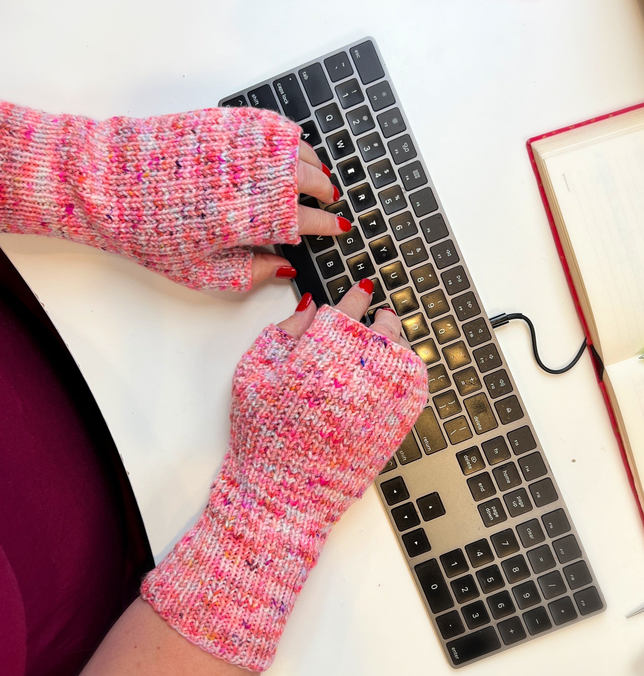 Hands wearing pink speckled knit fingerless mittens while typing on a keyboard beside an open notebook.