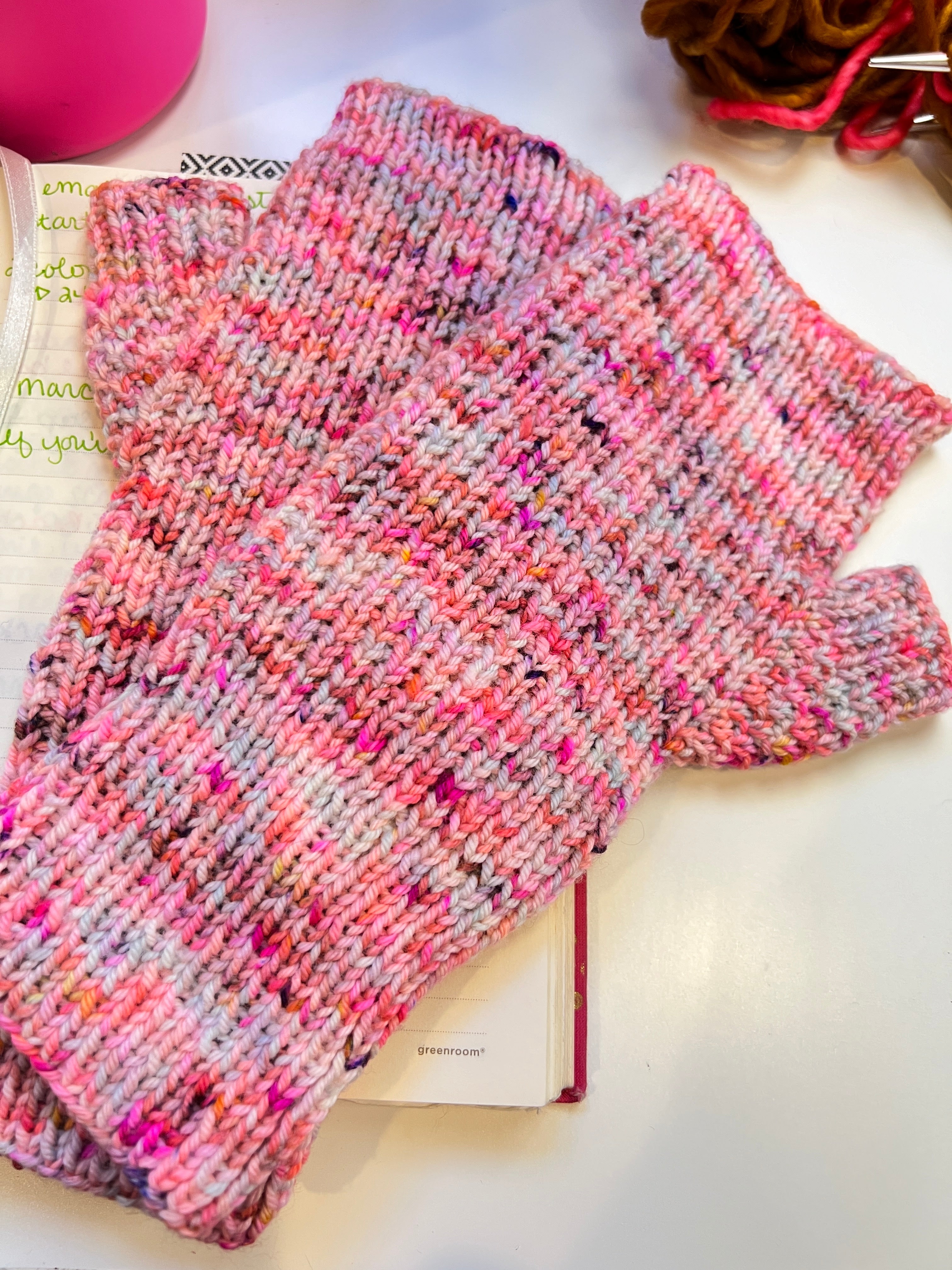 Pink and gray speckled fingerless mittens laid across an open notebook, showing broken rib texture and long ribbed cuffs.