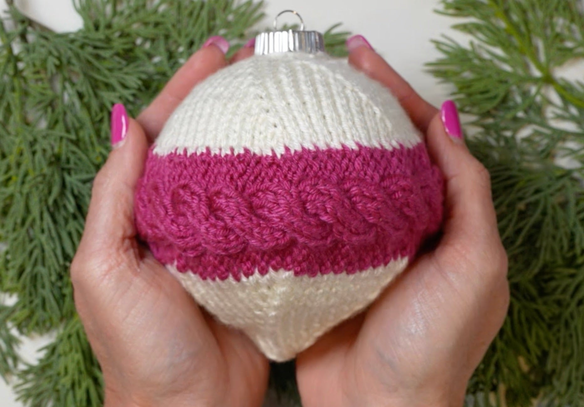 Two hands with pink nail polish hold a round ornament featuring the “My Tree Needed This Knit Ornament Pattern PDF – Knit Round Ornament Flat,” covered in charming white and pink cabled knitting inspired by Marly Bird. Green pine branches are visible in the background.