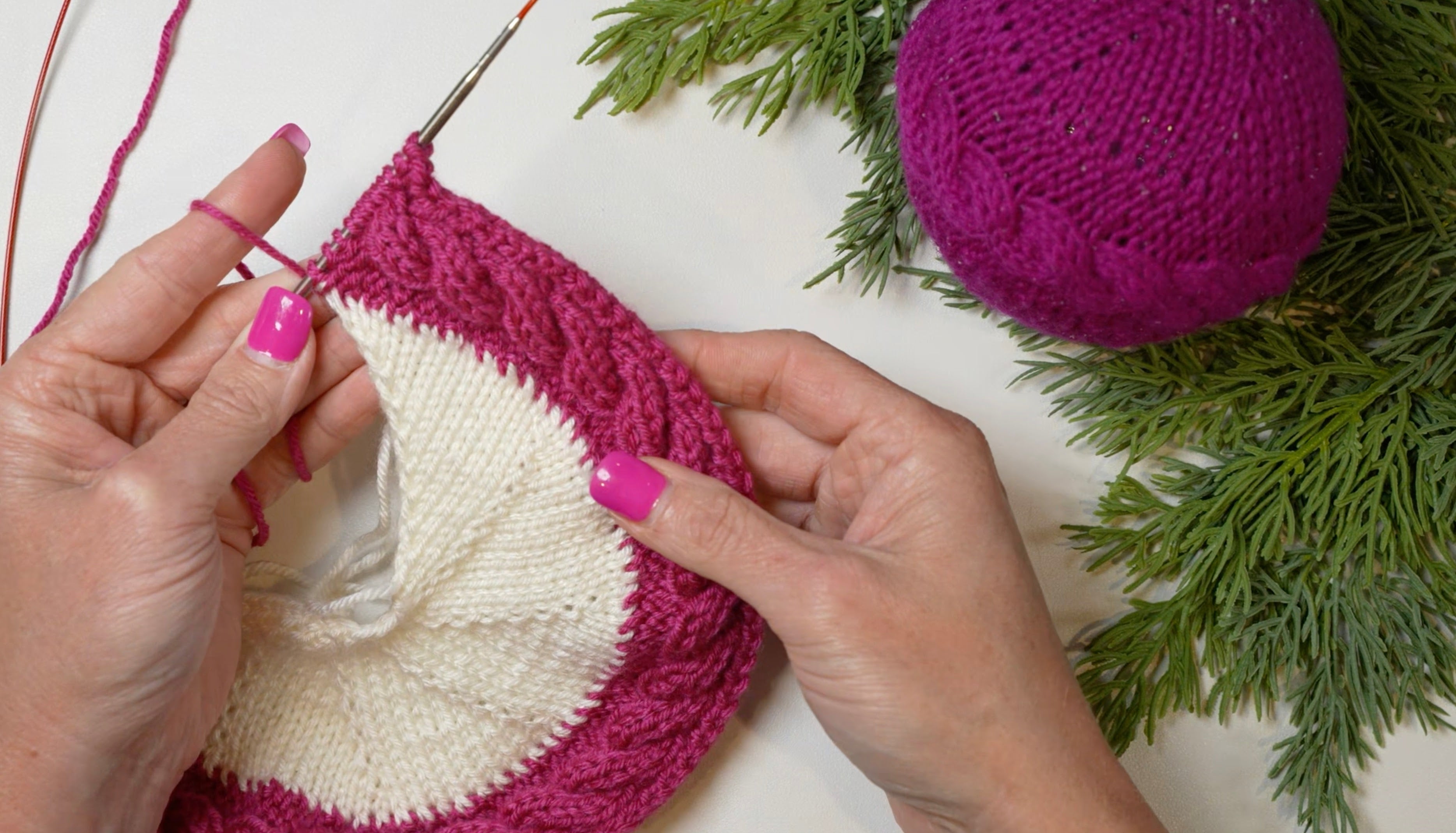 Hands with pink nail polish knitting a white and magenta circular piece, using the “My Tree Needed This Knit Ornament Pattern PDF - Knit Round Ornament Flat” with magenta yarn, next to a ball of yarn and green pine branches on a white surface.