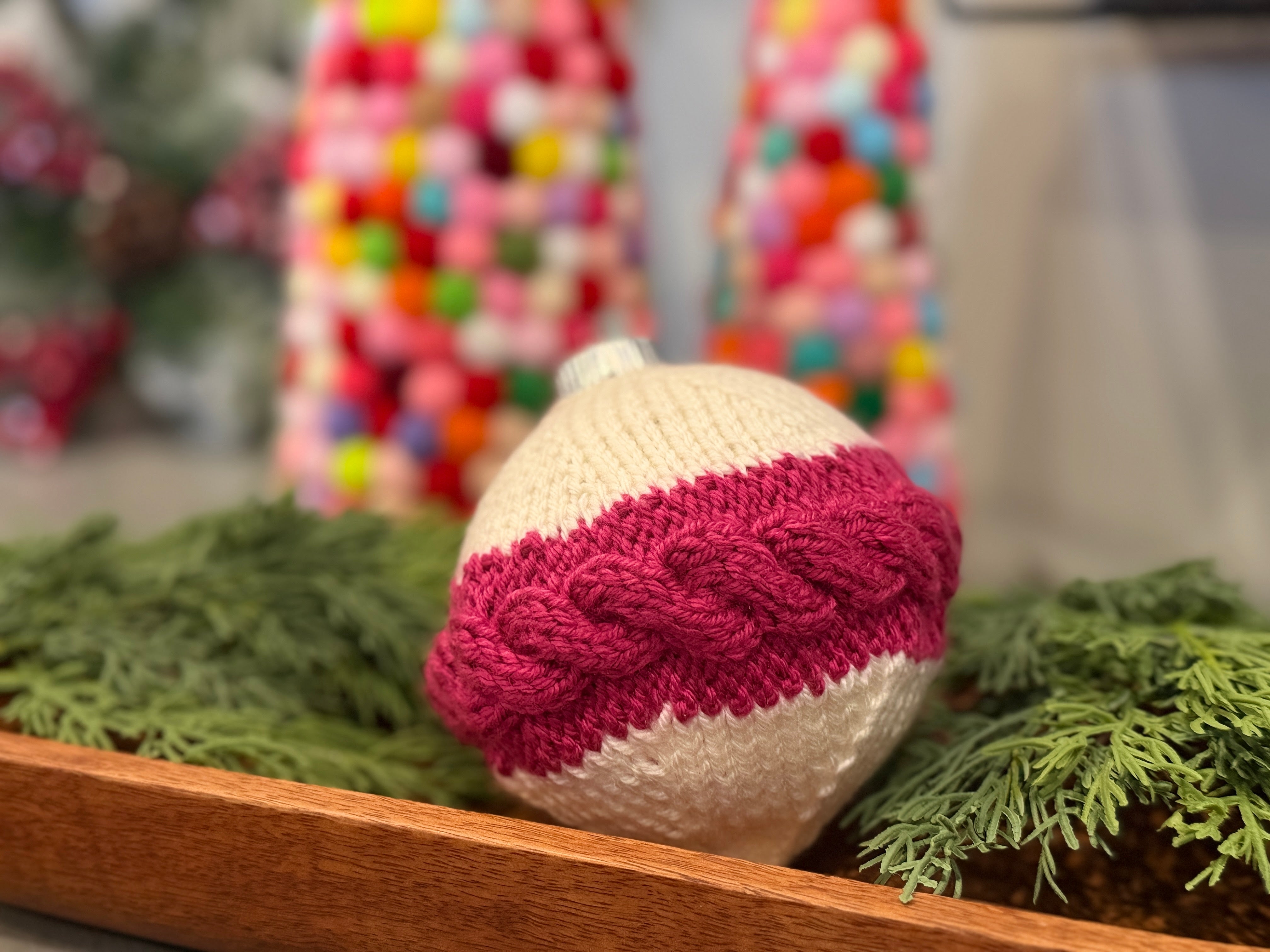 A festive white ornament, showcasing the “My Tree Needed This Knit Ornament Pattern PDF - Knit round ornament Flat” with a pink knitted braid inspired by a Marly Bird cabled cover, rests on greenery in a wooden tray while colorful, blurred decorations fill the background.