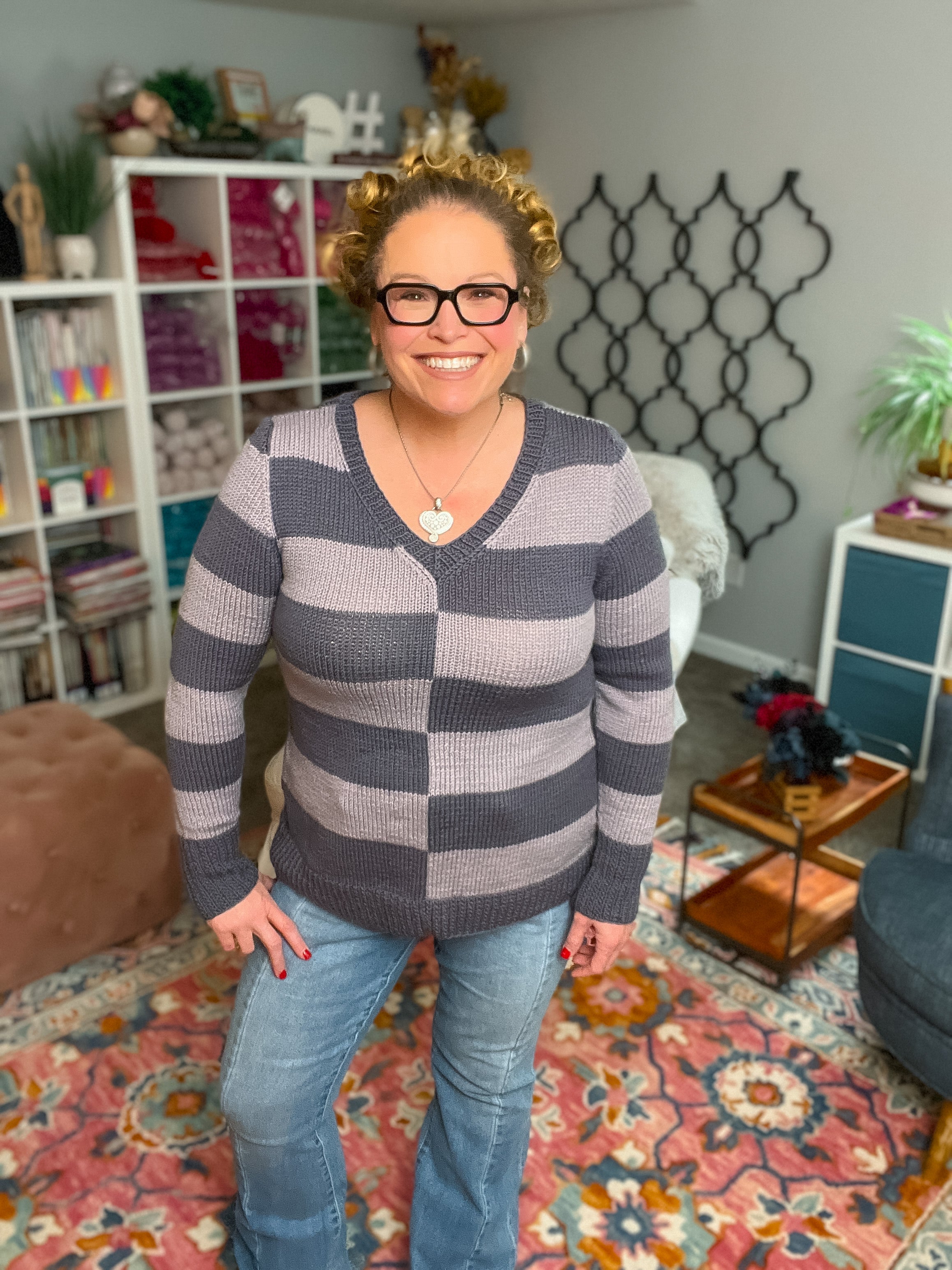 A smiling woman with curly hair and glasses stands in a cozy, colorful room wearing the My Favorite Knit Sweater Pattern. She's paired it with jeans, while bookshelves, decor, and a vibrant rug create a welcoming backdrop.