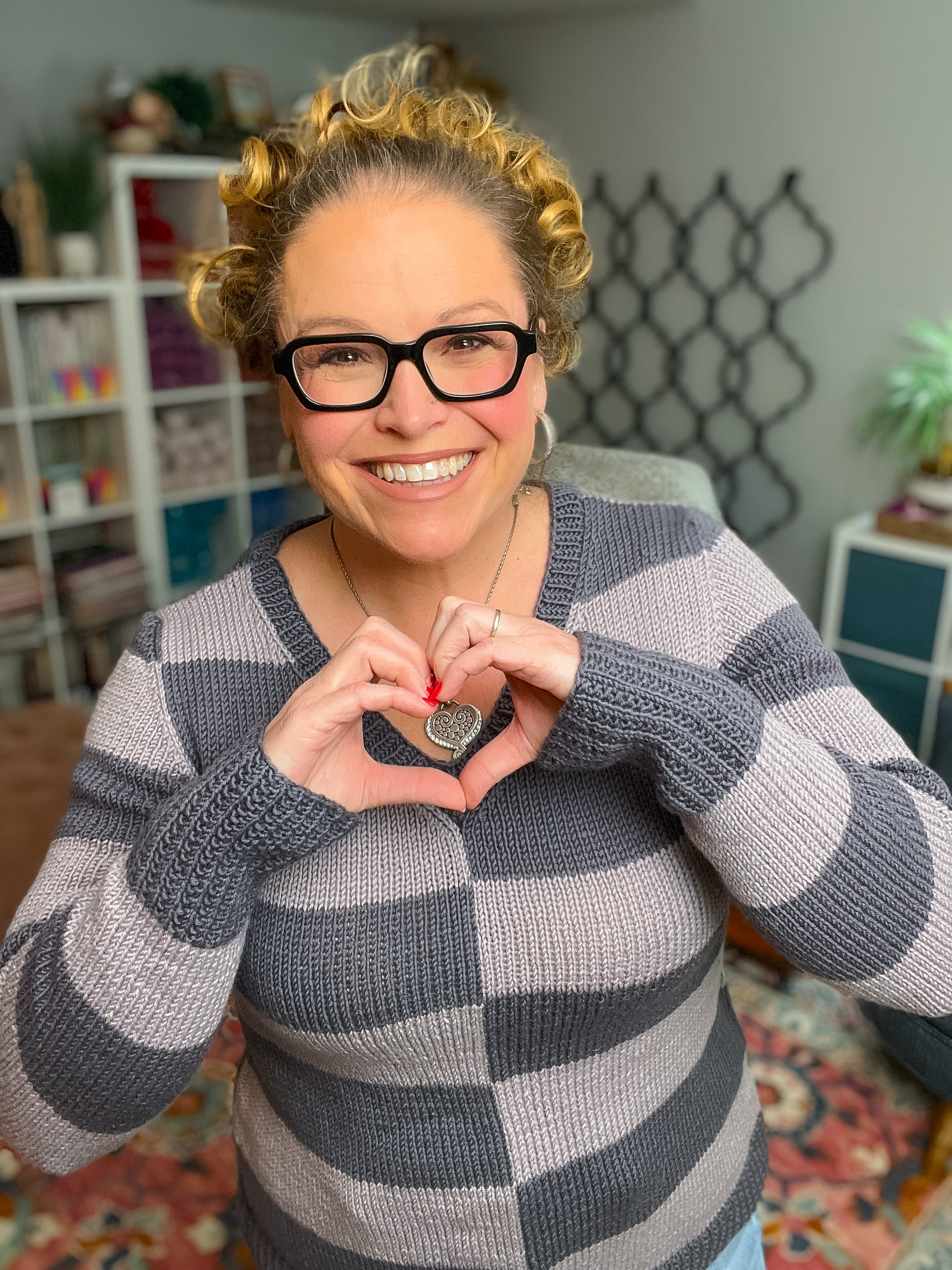 A smiling woman with curly hair and glasses wears the My Favorite Knit Sweater Pattern, forms a heart shape with her hands holding a heart pendant, standing in a cozy, colorful room filled with shelves and plants.