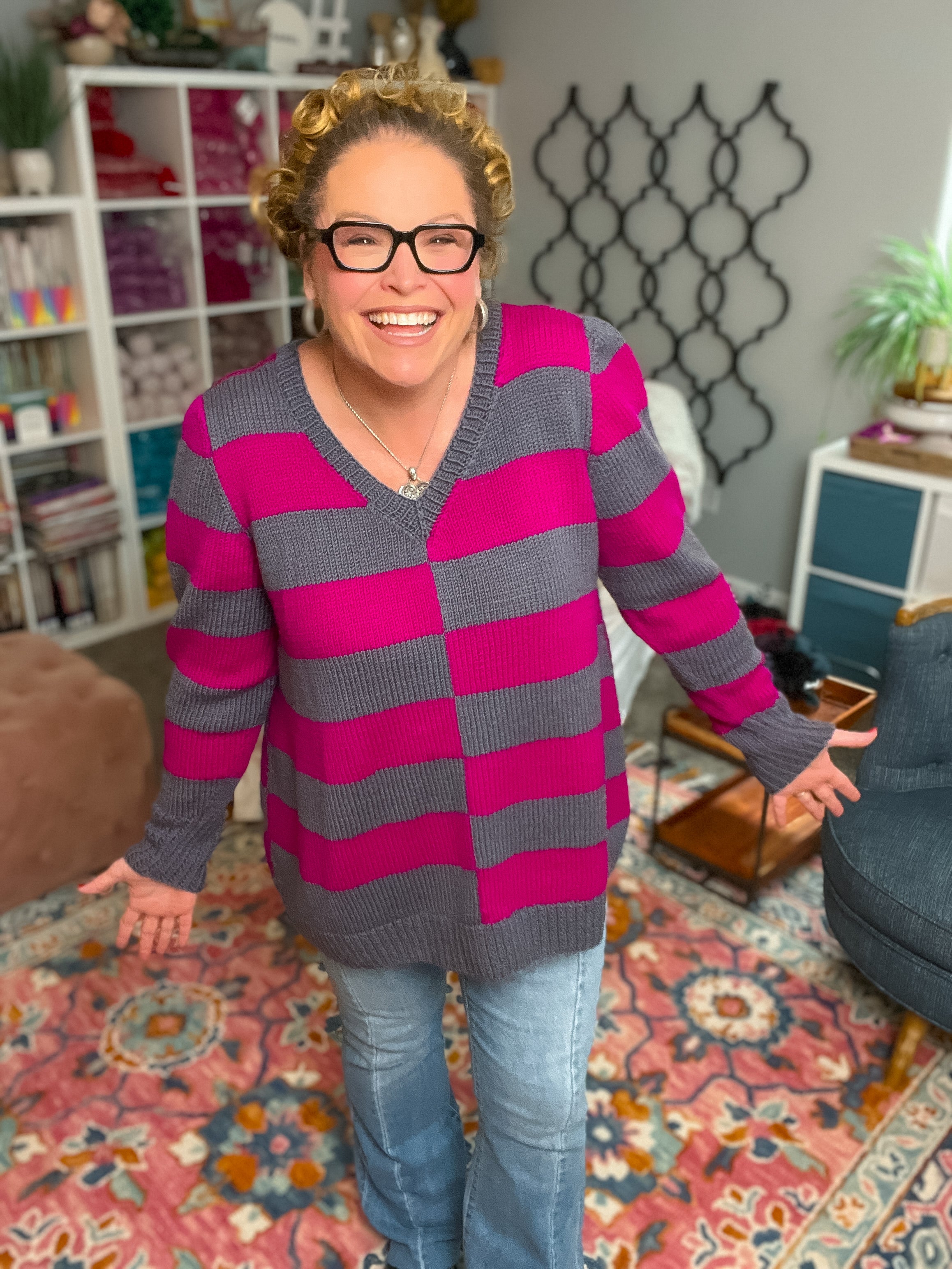 A smiling person with curly hair and glasses wears the My Favorite Knit Sweater Pattern in purple and magenta stripes, paired with jeans. They stand in a cozy, colorful room with bookshelves, a patterned rug, and home decor in the background.