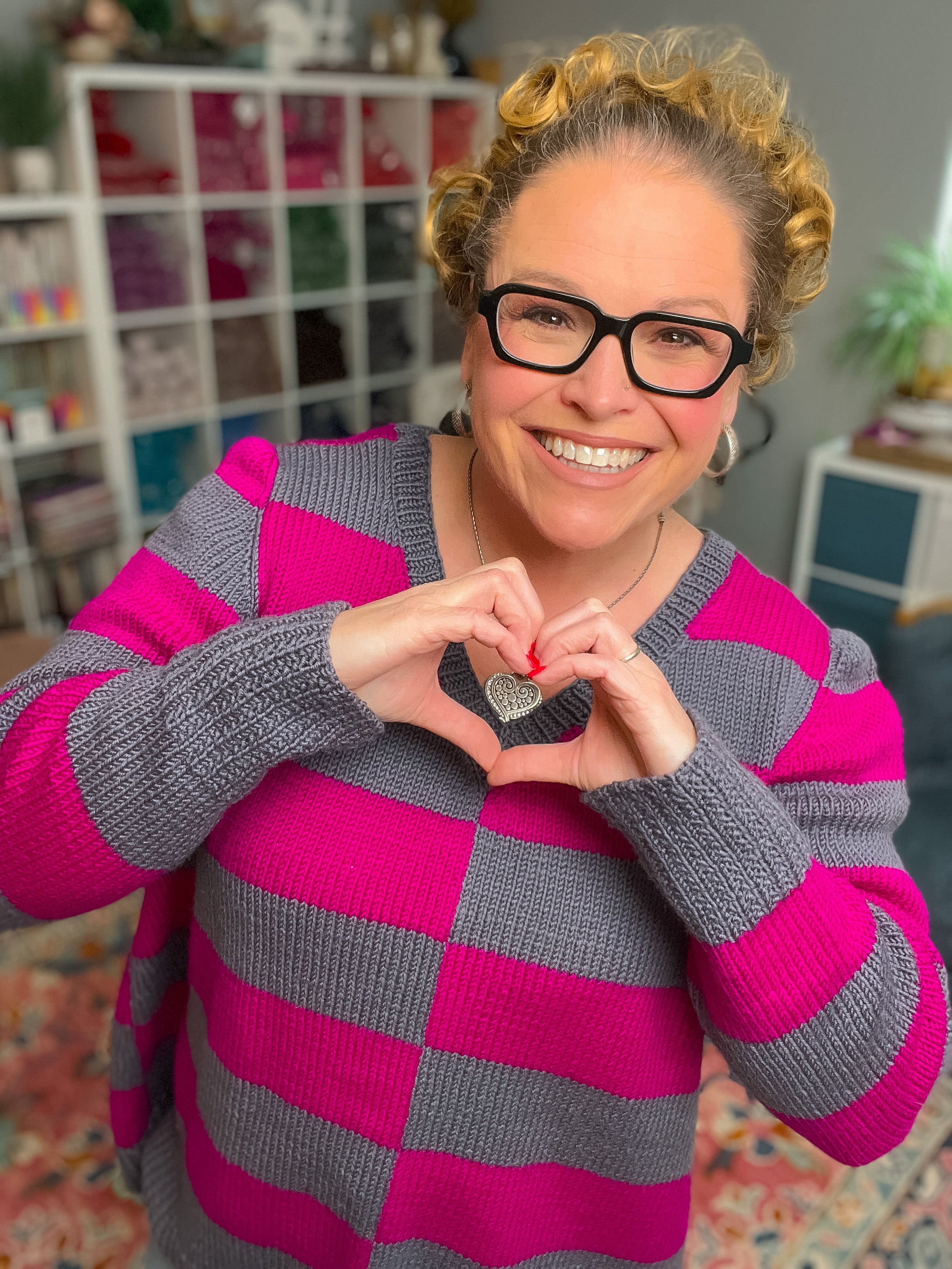A smiling person with curly hair and glasses models the My Favorite Knit Sweater Pattern—a pink and gray striped sweater—while forming a heart shape with their hands in front of a colorful indoor shelf.