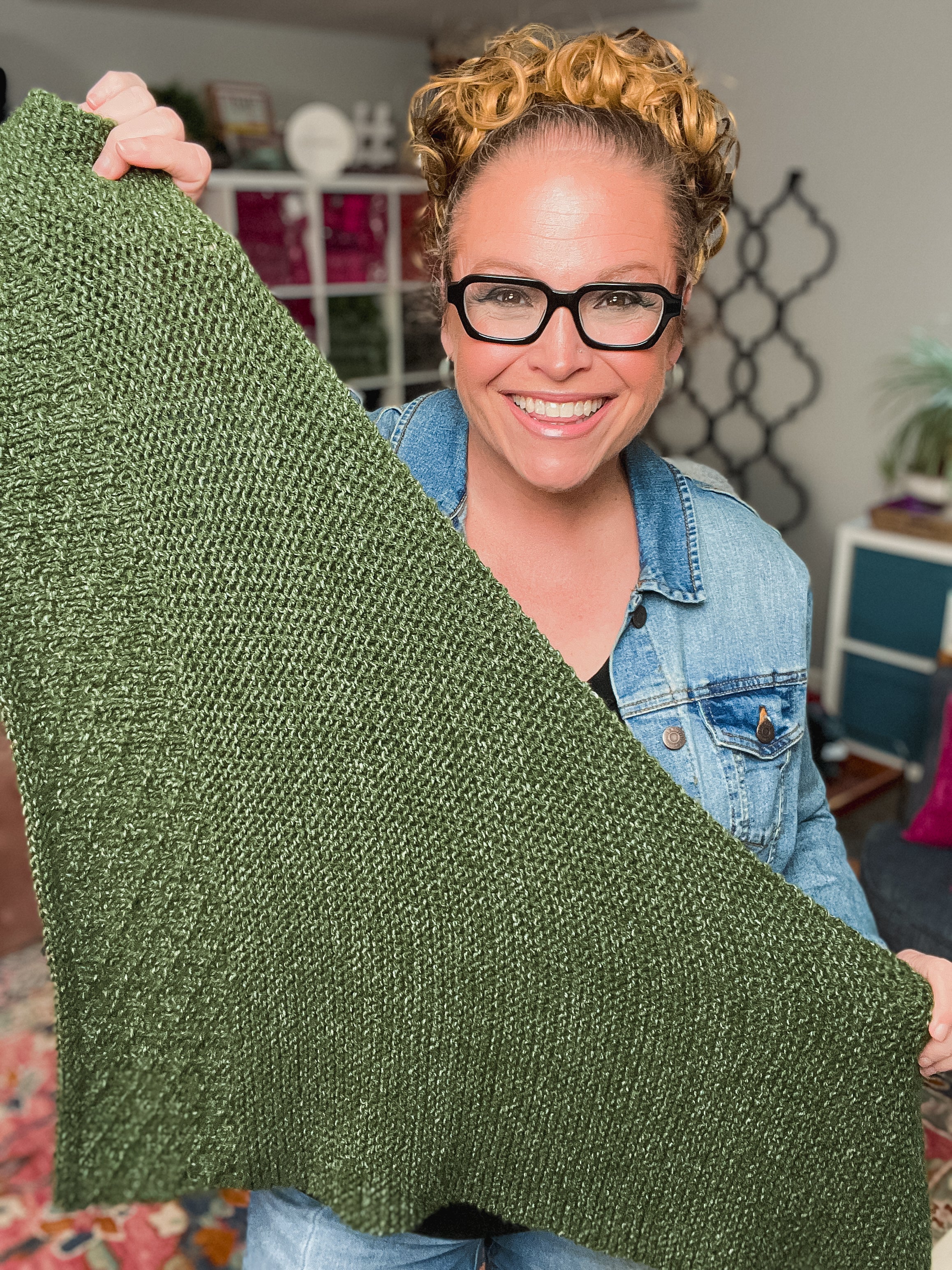A smiling woman with glasses and curly hair holds up a textured green knit blanket. She's wearing a denim jacket in a cozy room—perfect inspiration for the Peak Serenity Knit Shawl Pattern by Marly Bird, ideal for beginner knitters.