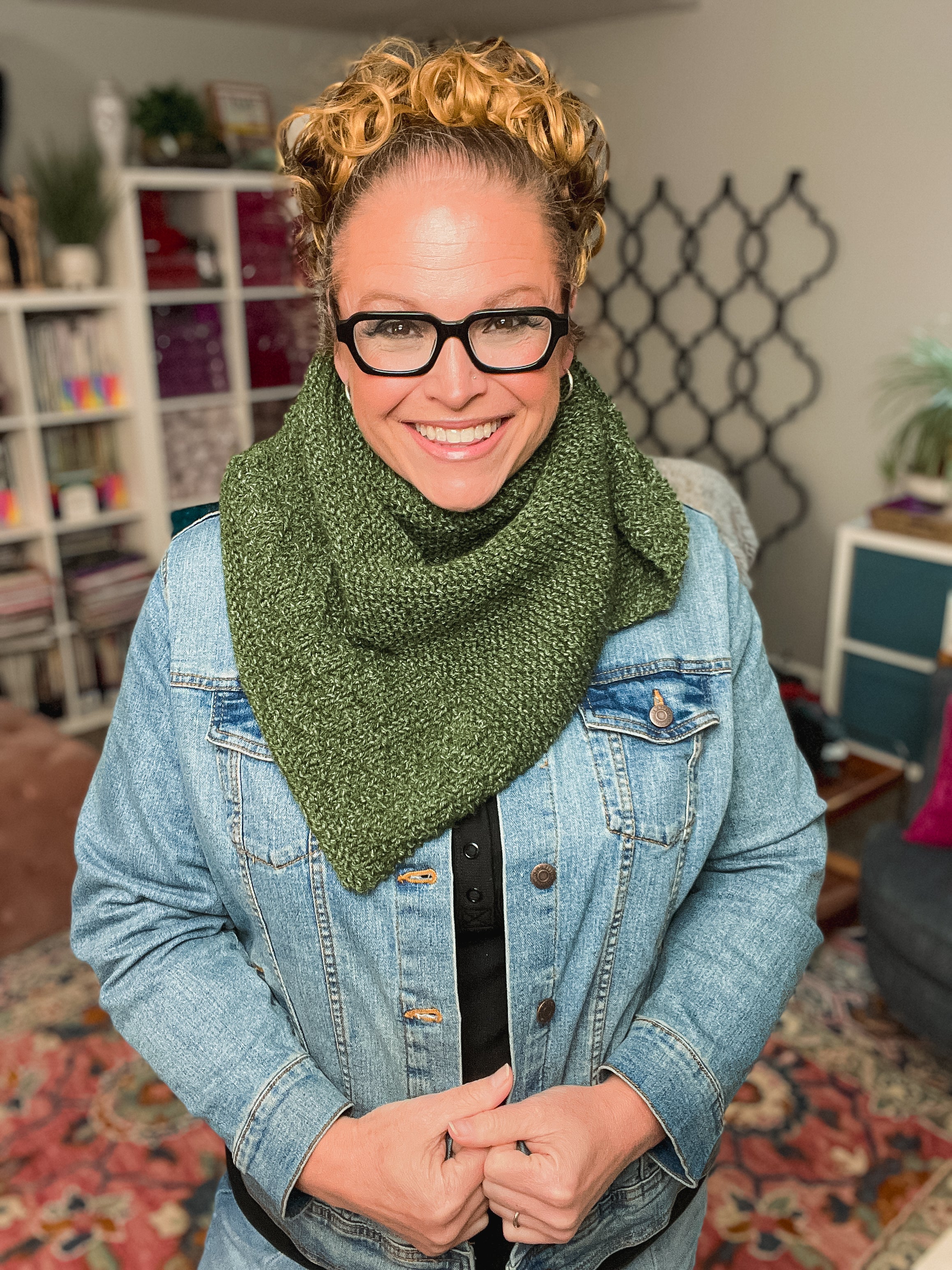 A smiling woman with curly hair and glasses models the Peak Serenity Knit Shawl, a beginner-friendly knitting pattern, styled with a denim jacket. She stands indoors, surrounded by shelves, books, and plants.