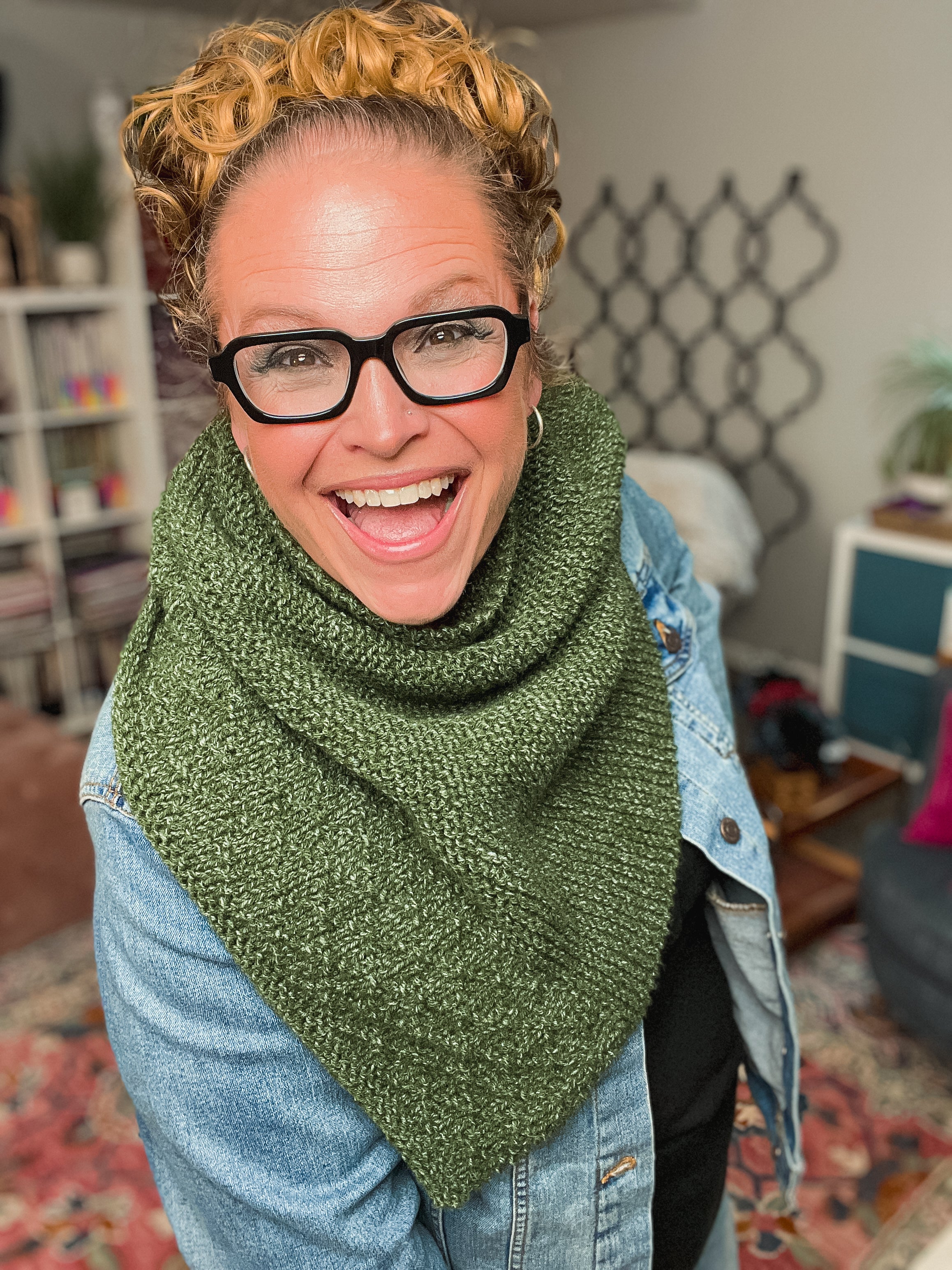 A smiling person with glasses and curly hair wears a green knit scarf made using the Peak Serenity Knit Shawl Pattern, paired with a denim jacket, standing indoors in a cozy room filled with shelves and plants.