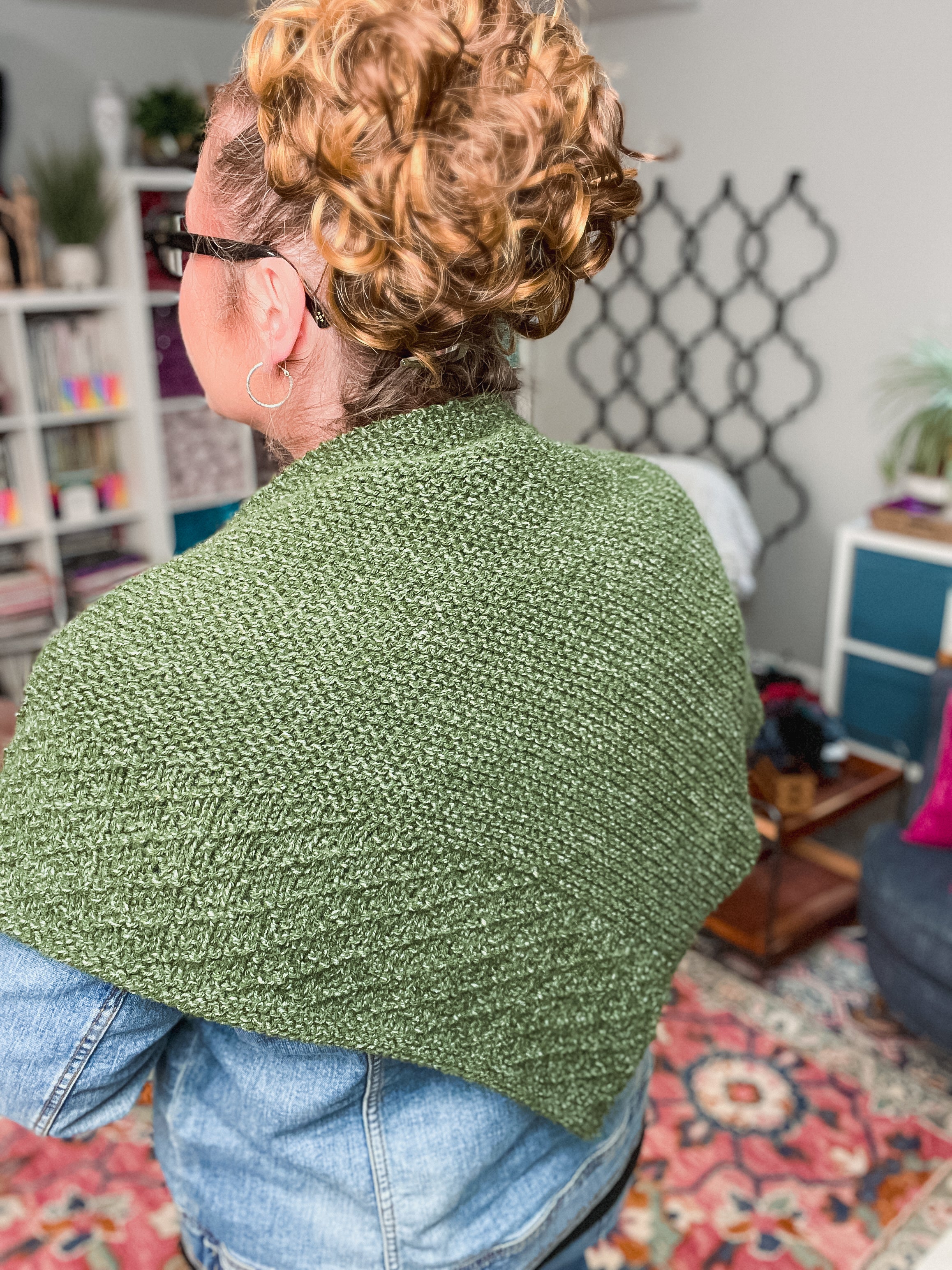 A person with curly hair in a bun, glasses, and a denim jacket wears the Peak Serenity Knit Shawl Pattern in green, standing in a cozy room with bookshelves, plants, and colorful decor in the background.