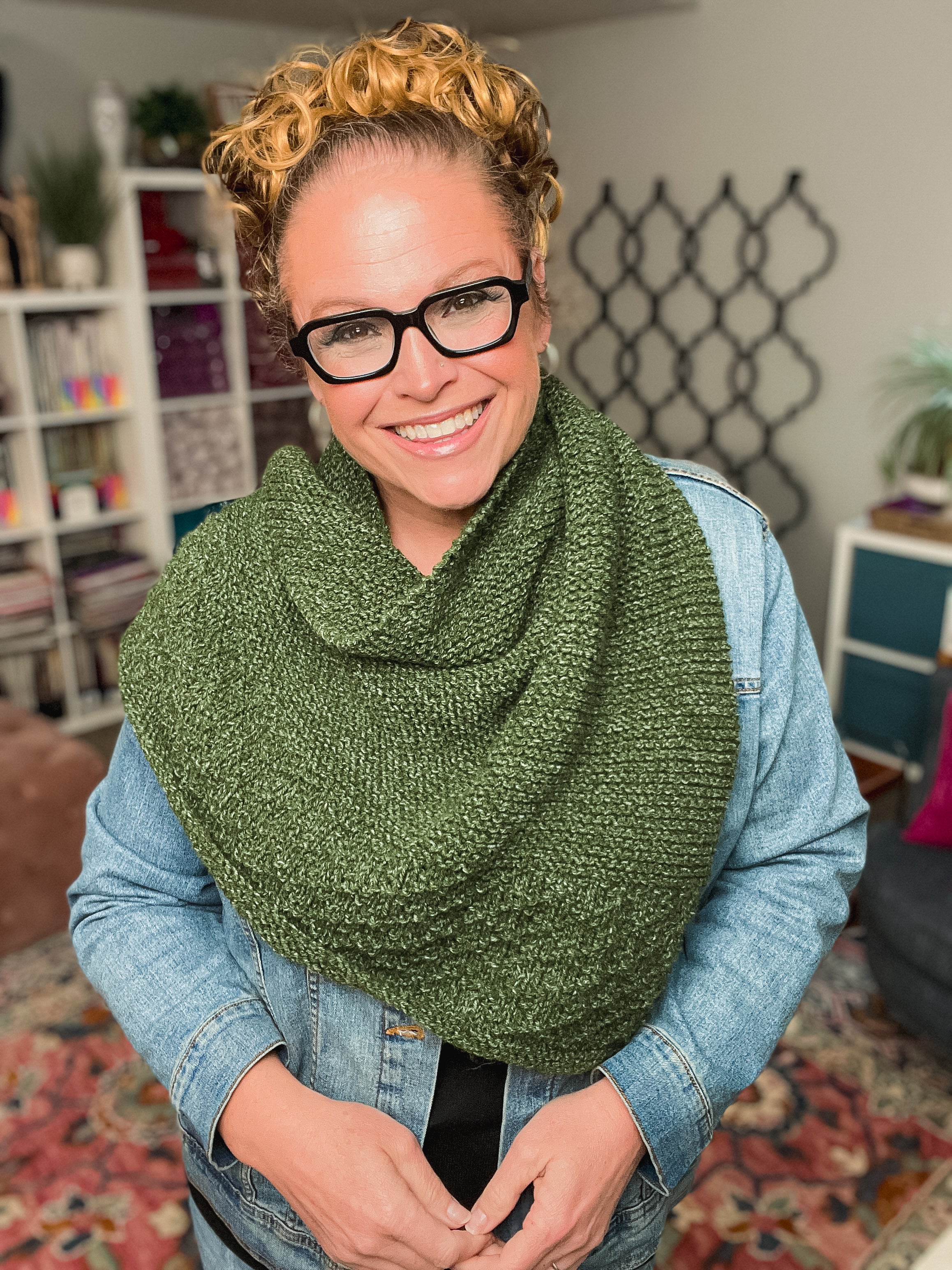 A smiling woman with curly blonde hair and glasses wears a green shawl made using the Peak Serenity Knit Shawl Pattern and a denim jacket, standing in a cozy room with bookshelves and patterned decor in the background.