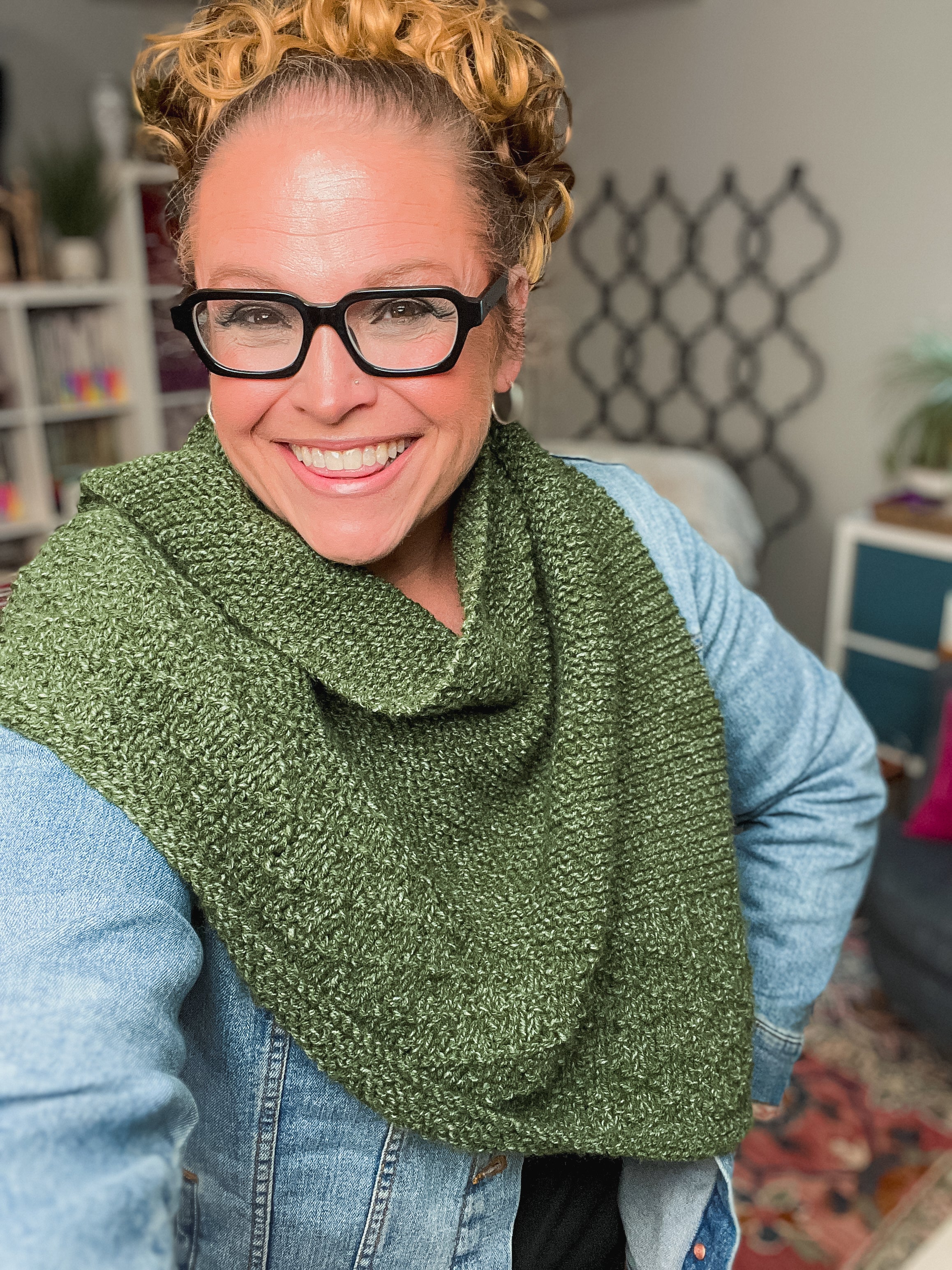 A woman with curly hair and black glasses, wearing a denim jacket and the Peak Serenity Knit Shawl Pattern in green, smiles at the camera in a cozy room with colorful bookshelves and patterned decor in the background.