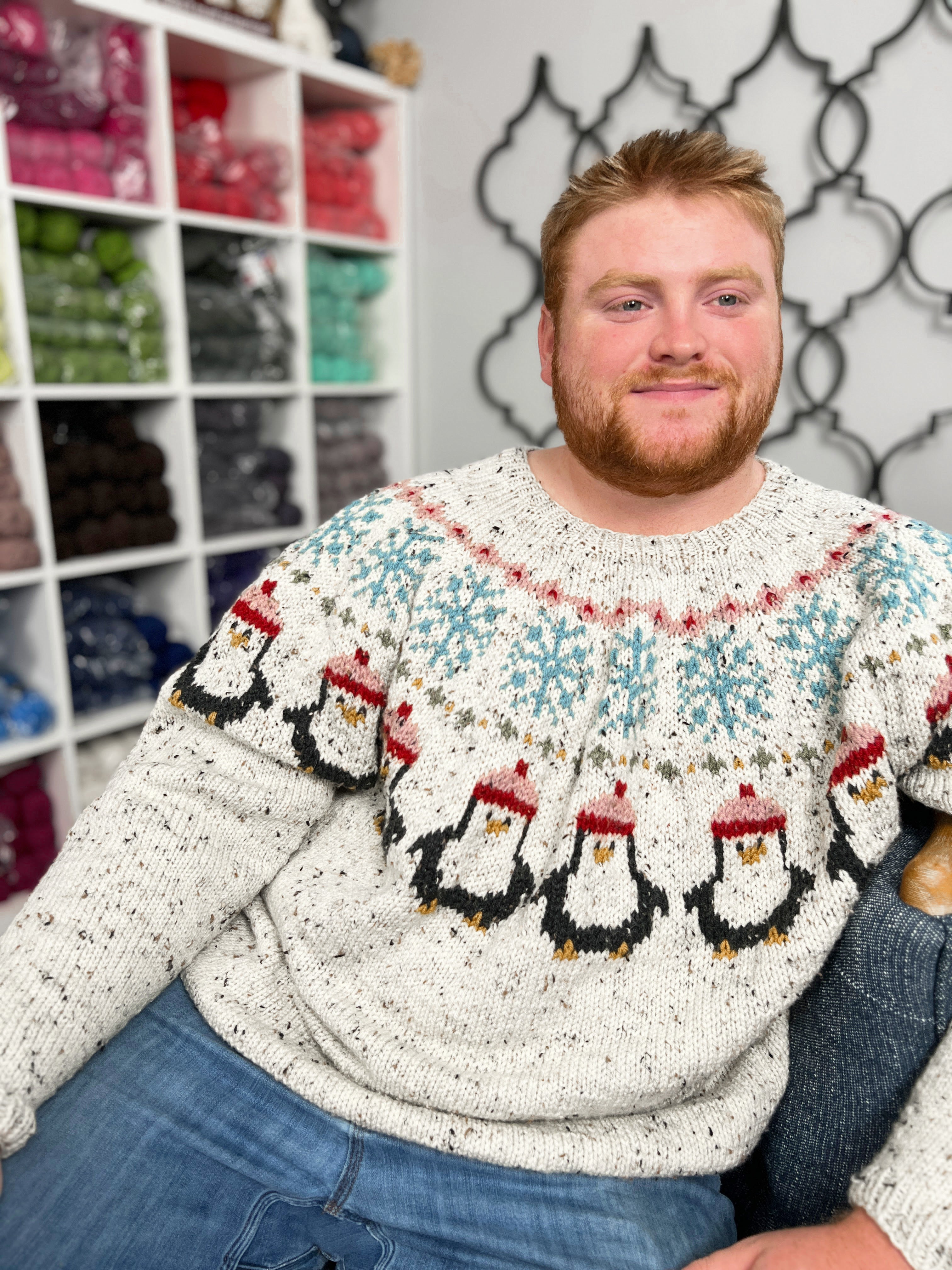 Man wearing a sweater with penguin design in a room with shelves and patterned wall.