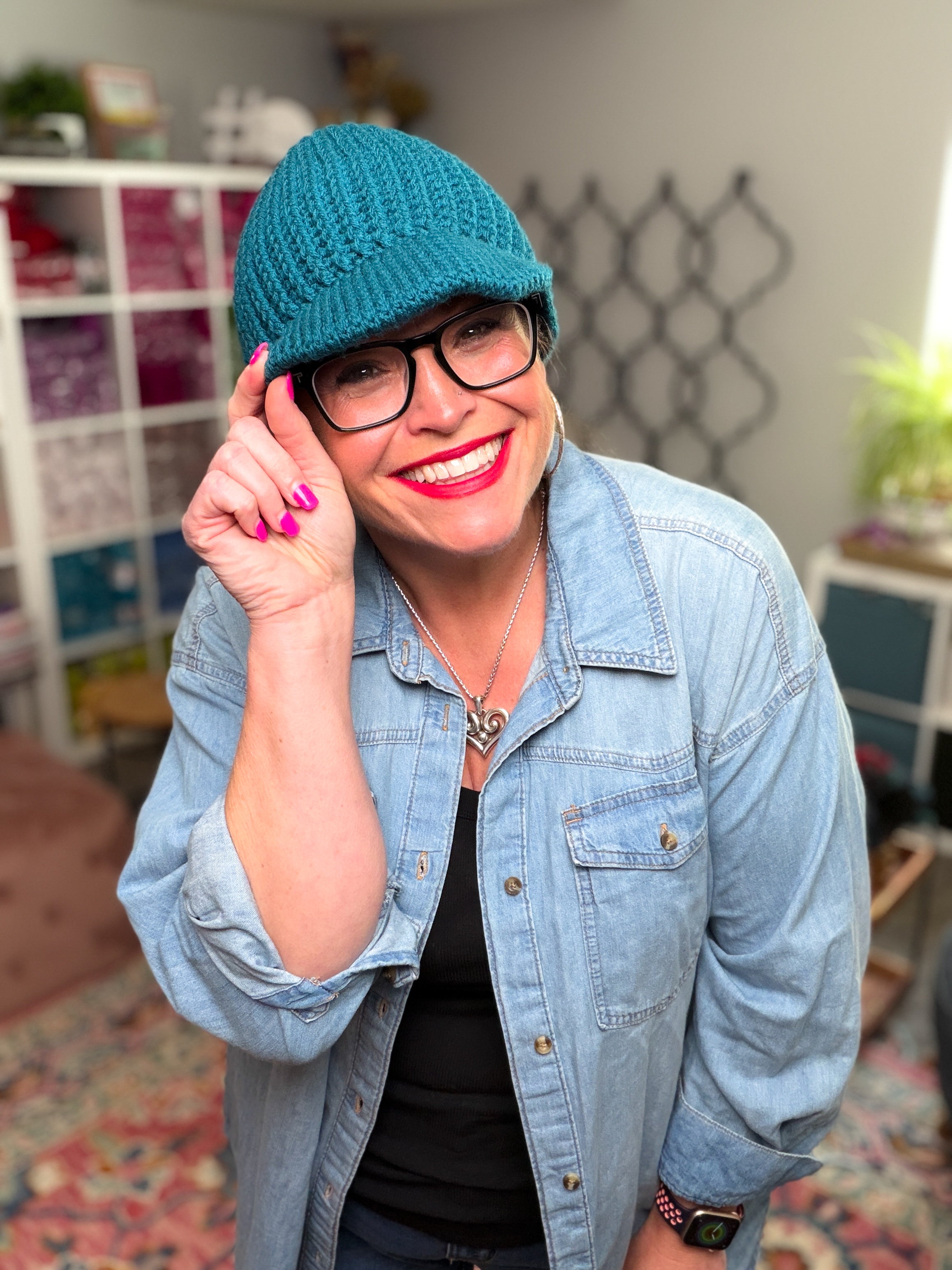 A smiling person in glasses models the “Ribbed for Him & Her Crochet Hat,” a teal ribbed design, while wearing a denim shirt and adjusting the hat with one hand. In the background, colorful shelves and a patterned rug add to the cozy scene.