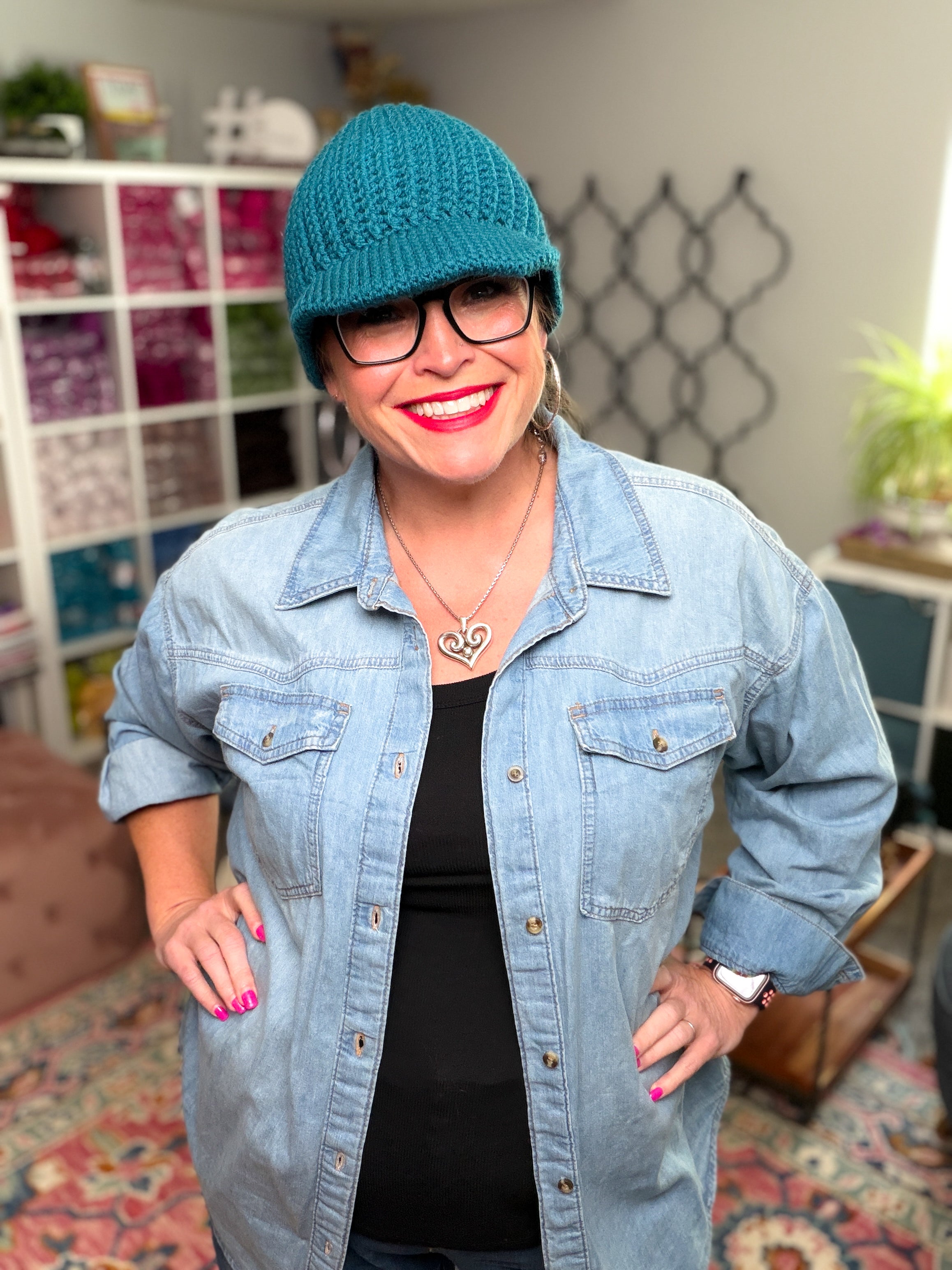 A smiling woman models the Ribbed for Him & Her Crochet Hat (pattern PDF) in teal, paired with glasses, a denim shirt, and a black top as she stands in a colorful room filled with yarn shelves, a patterned rug, and indoor plants.