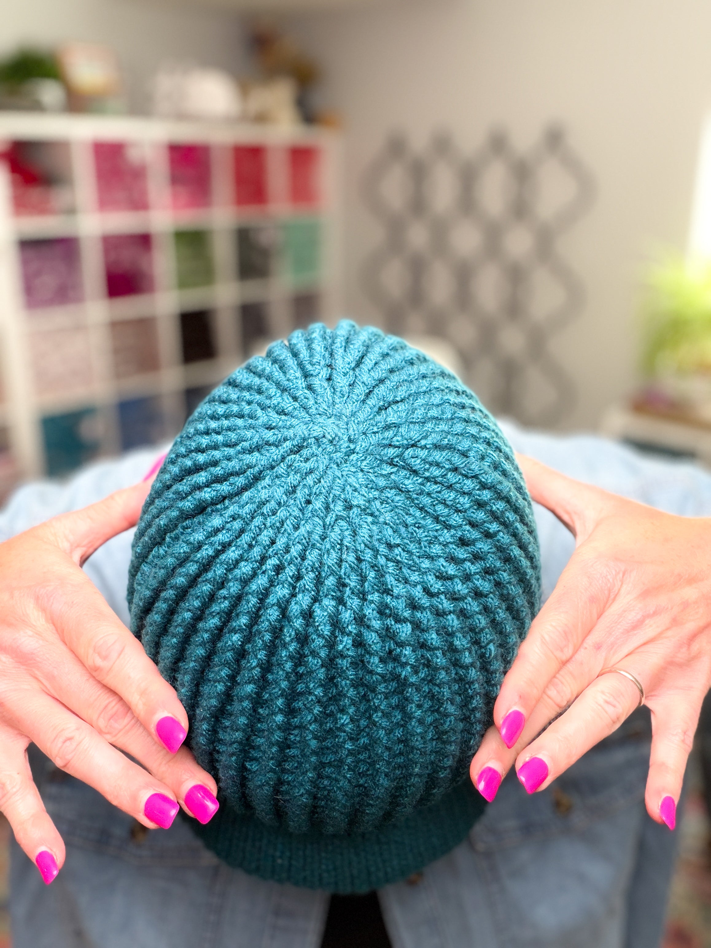 Hands with bright pink nail polish hold their head, showcasing the top of a teal Ribbed for Him & Her Crochet Hat (Pattern PDF). The blurred background features shelves filled with colorful yarn.