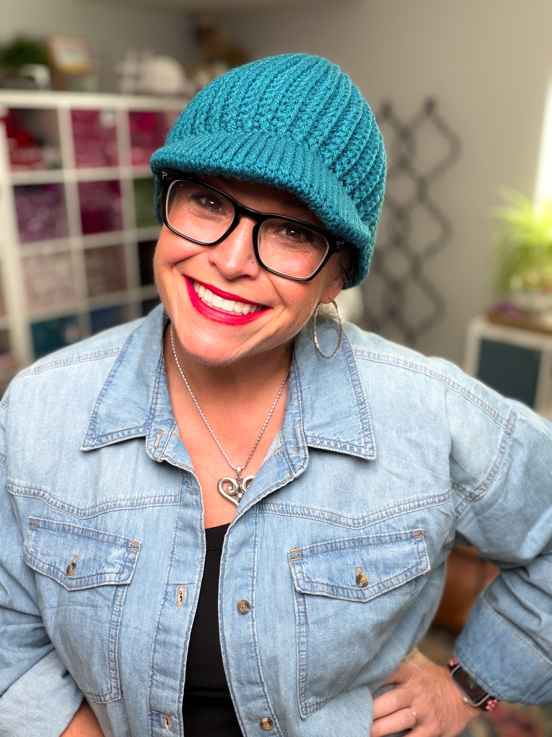 A smiling woman wearing glasses, hoop earrings, and a denim jacket stands indoors with shelves and plants in the background, modeling the Ribbed for Him & Her Crochet Hat Pattern PDF.