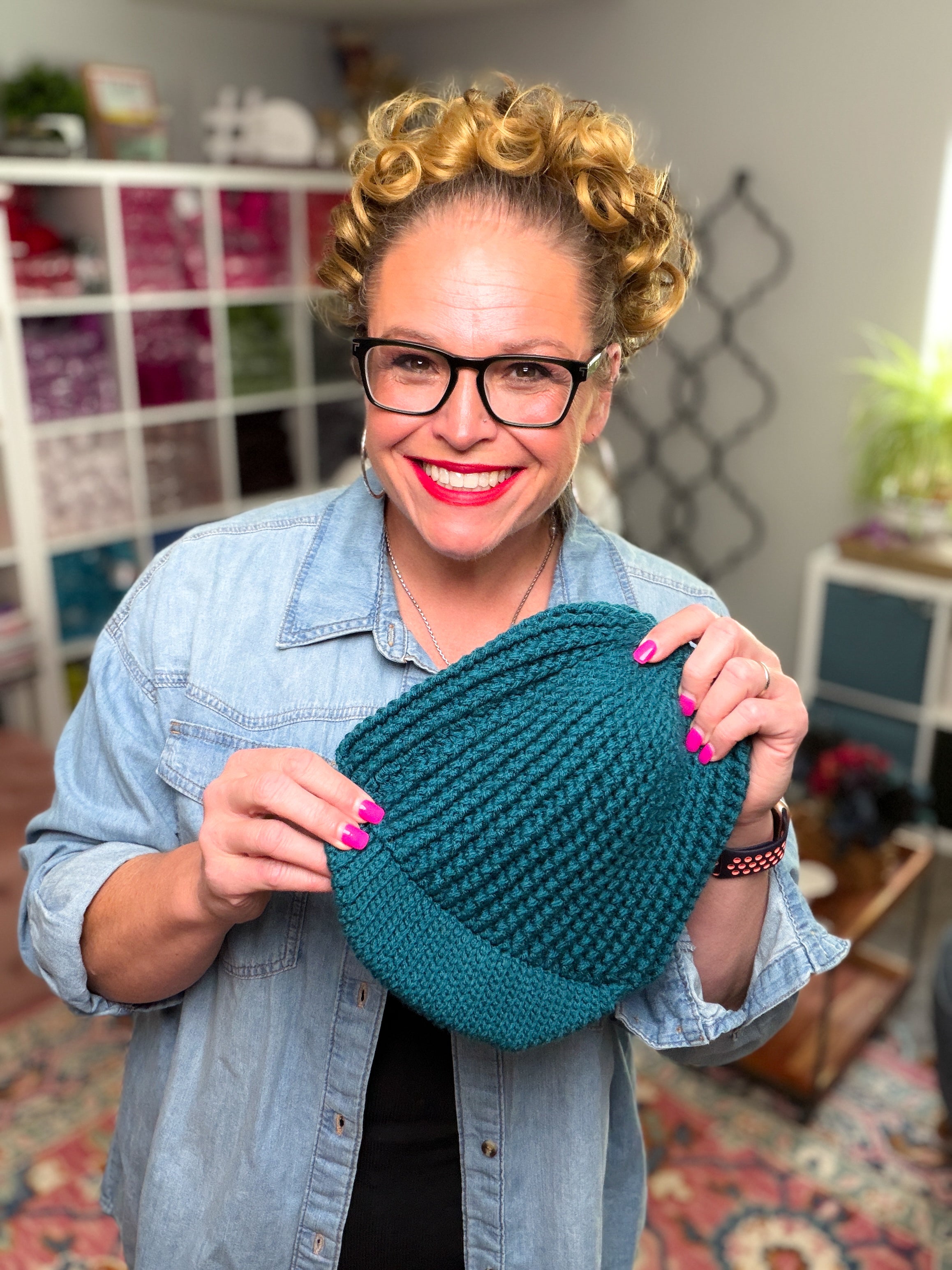 A smiling woman with curly hair and glasses holds the “Ribbed for Him & Her Crochet Hat Pattern PDF” in a teal ribbed crochet hat. She wears a denim shirt and bright pink nail polish, with shelves of yarn and a plant visible in the background.
