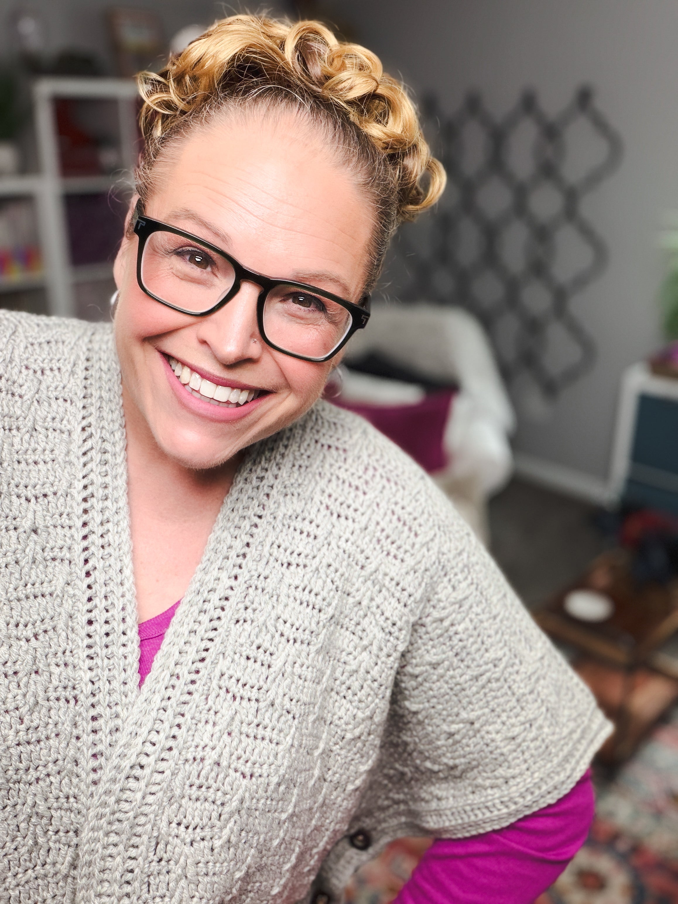 A woman with glasses and curly blonde hair smiles at the camera, modeling a cozy gray cardigan made with the Rockford Crochet Ruana Pattern over a pink top, standing in a warmly lit room with inviting home decor in the background.