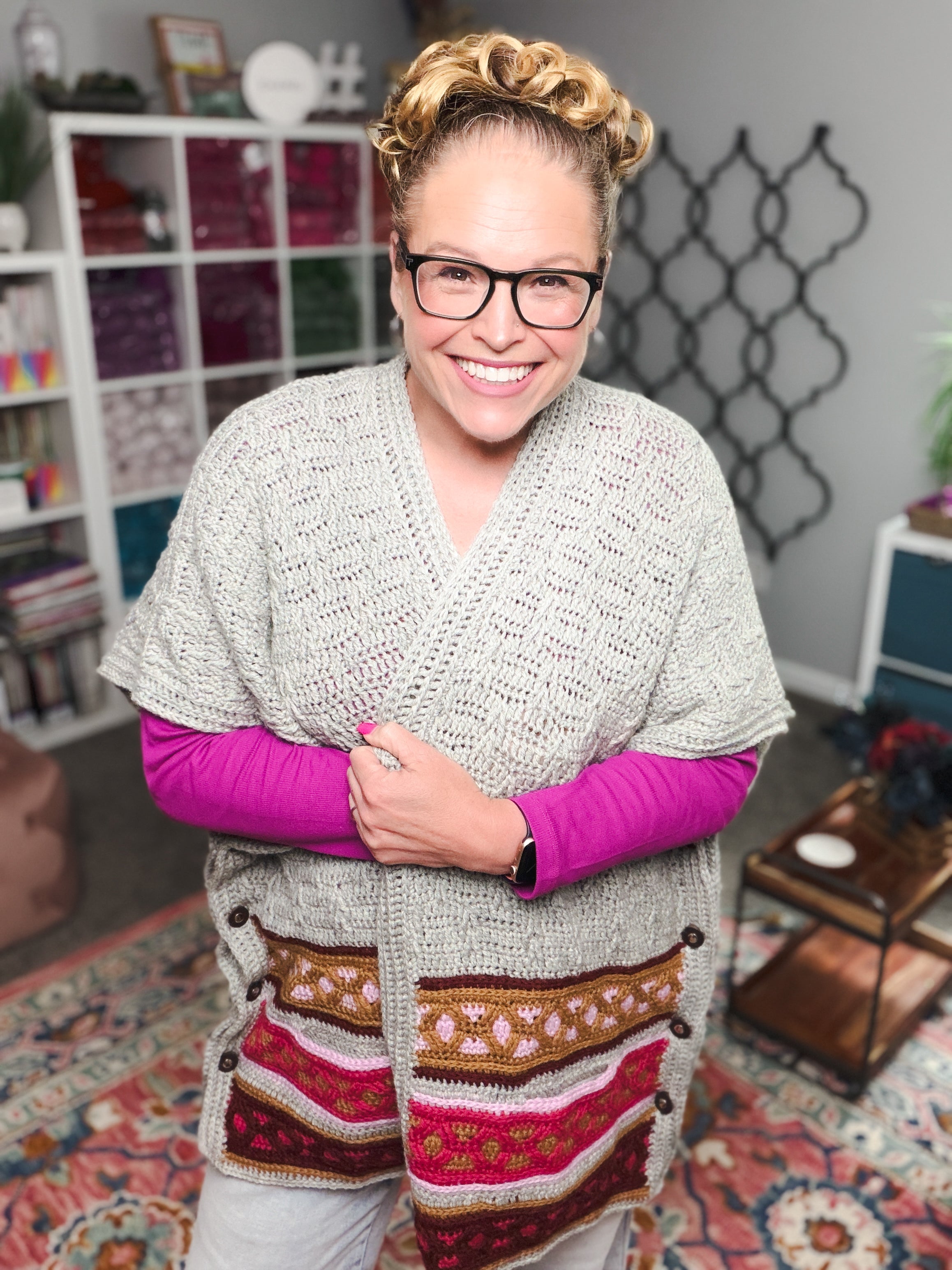 A smiling person with glasses and curly hair stands indoors, wearing a gray knit cardigan featuring the vibrant, mosaic-inspired design of the Rockford Crochet Ruana Pattern over a bright pink shirt. Shelves with yarn and a coffee table are visible in the cozy, well-lit room.