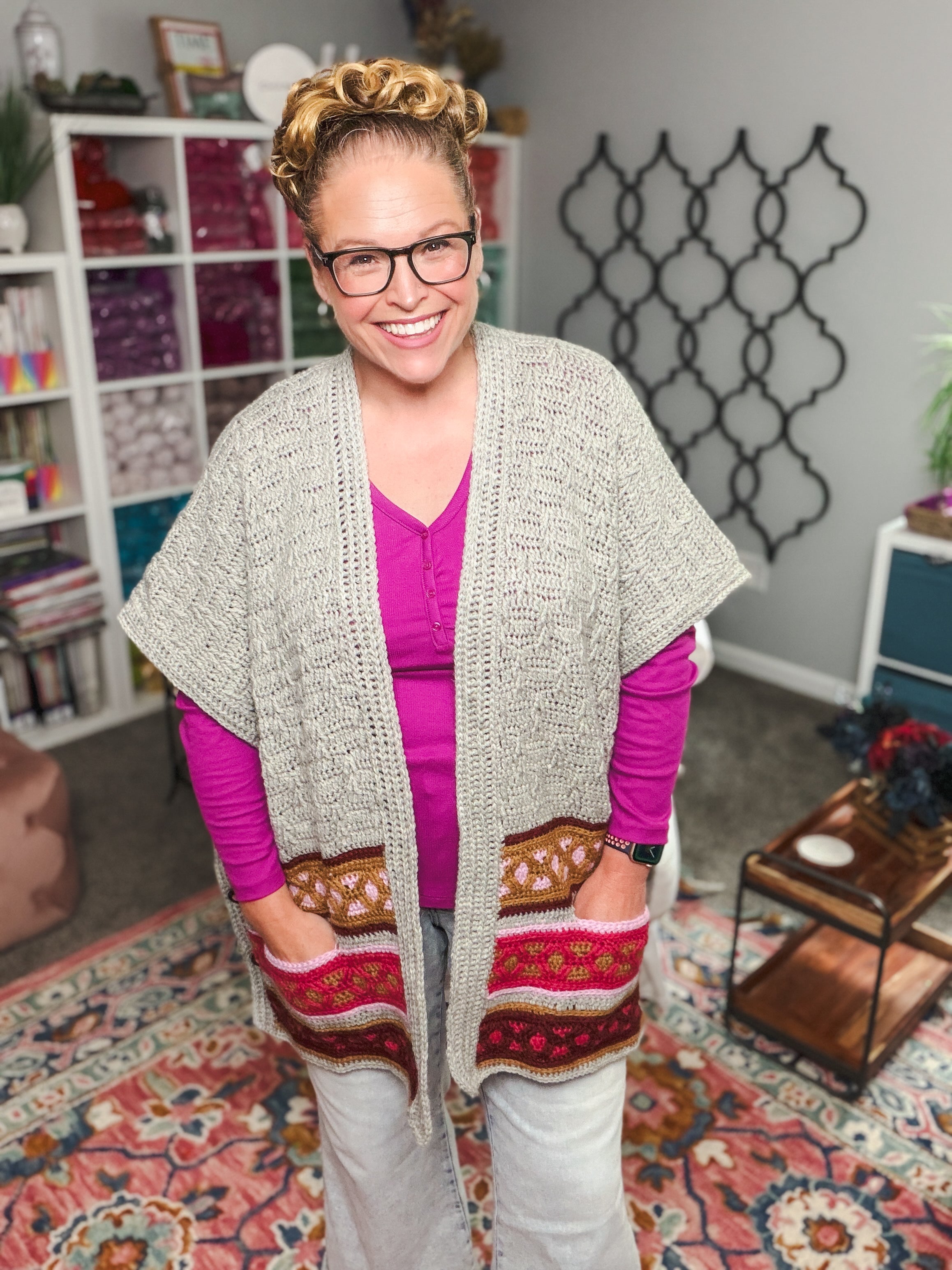 A smiling person with glasses wears the Rockford Crochet Ruana, featuring colorful pockets, over a pink shirt while standing in a cozy, vibrant room filled with yarn shelves and patterned decor.